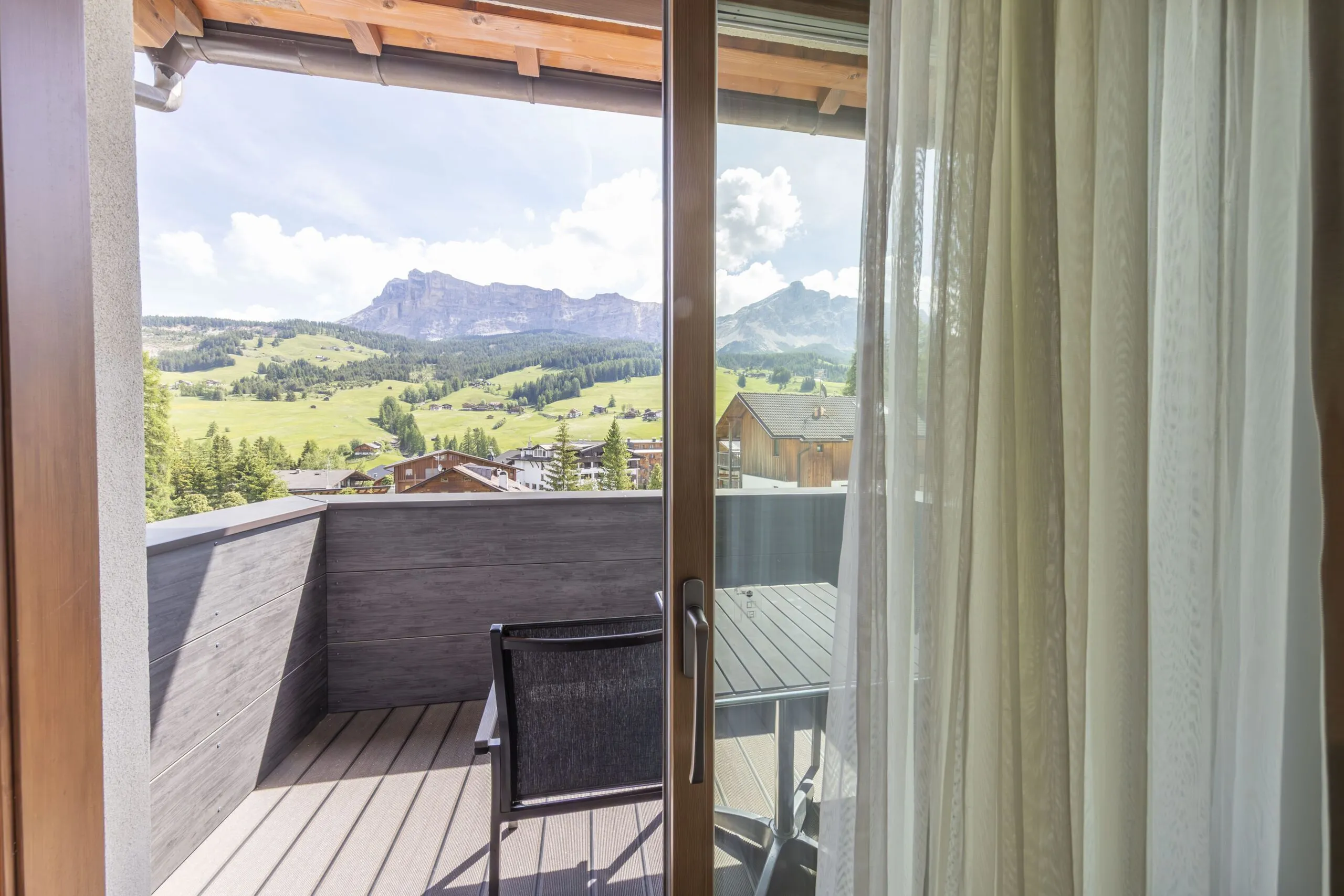 Balcony with wooden railing overlooking green hills, scattered houses, and distant mountains under blue sky.