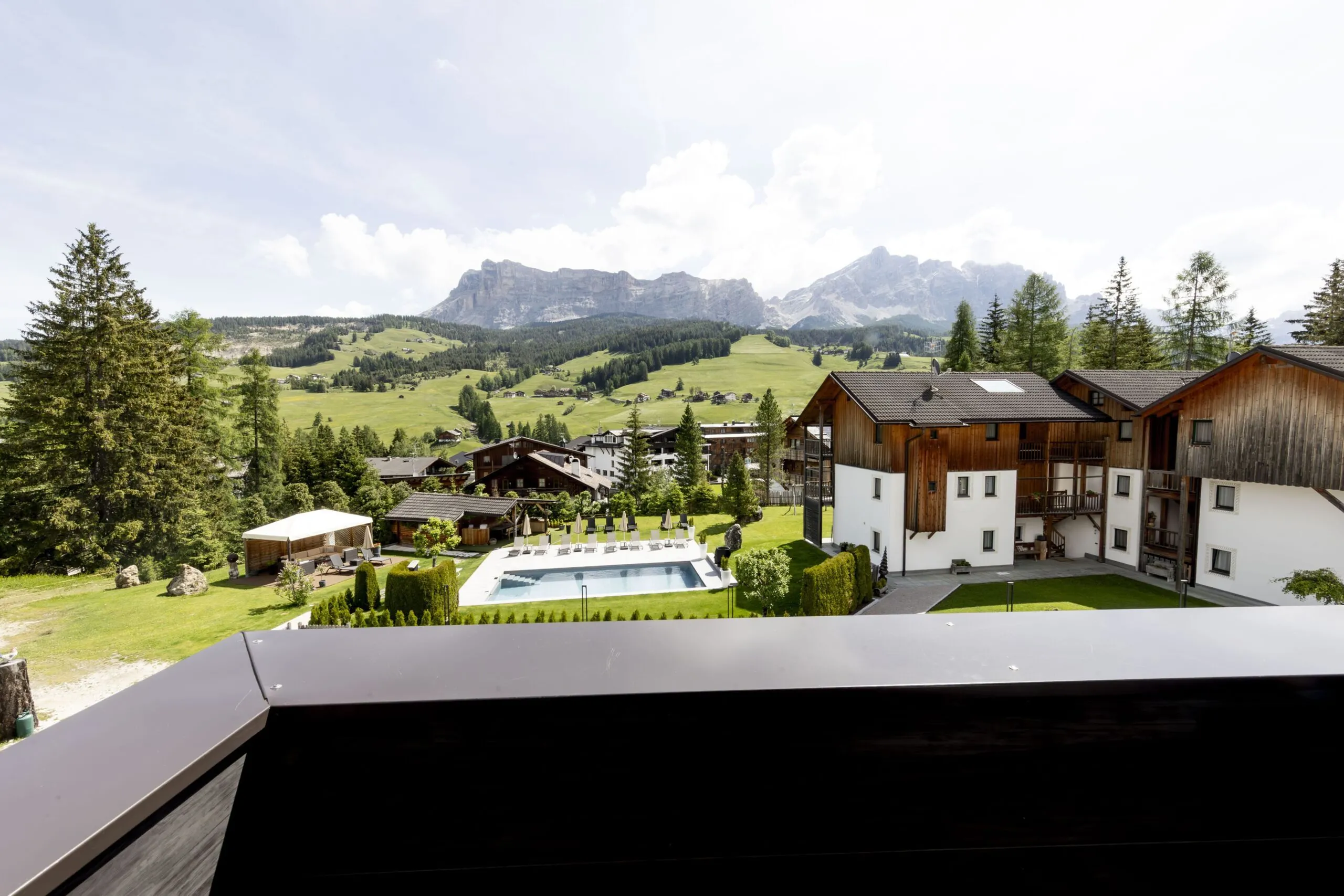 Mountain village with wooden chalets, pool, and green valley under distant mountains.