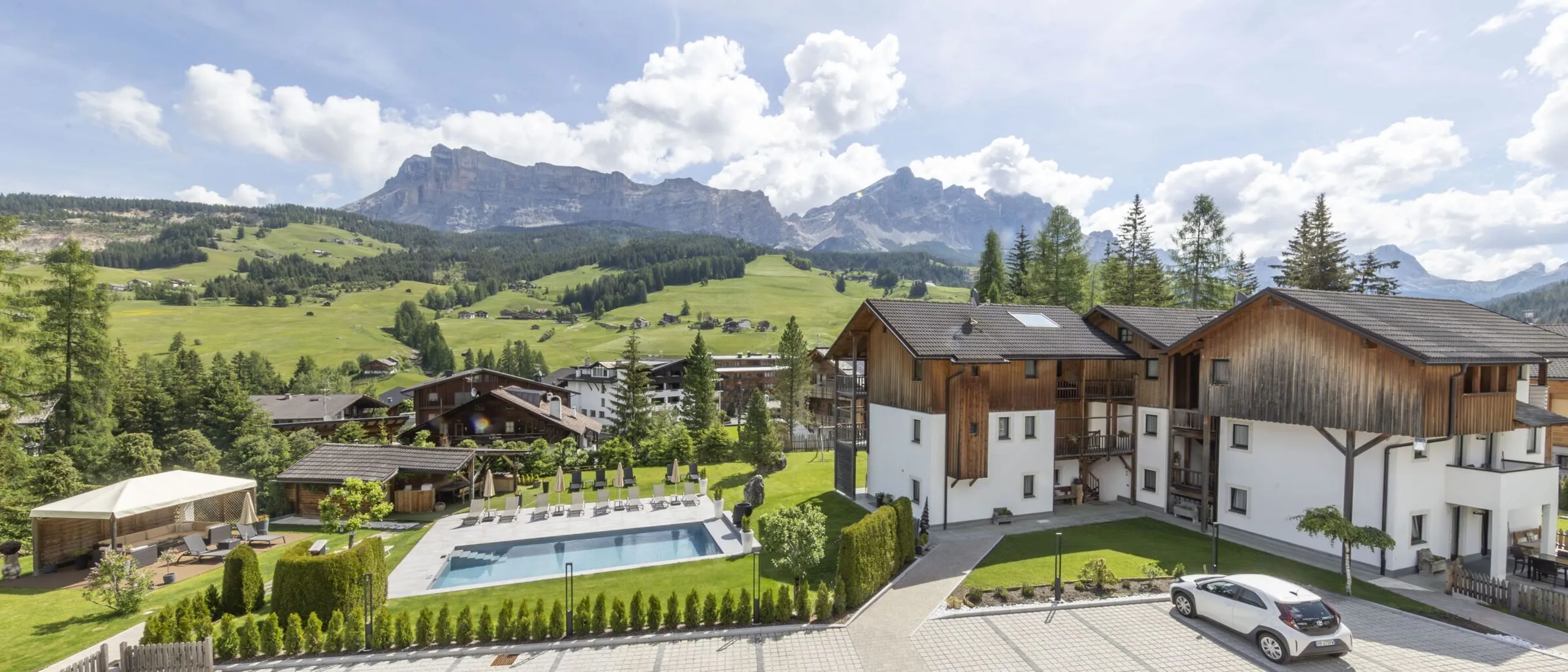 Charming alpine lodge complex with wooden facades, a swimming pool, and green fields under mountains.