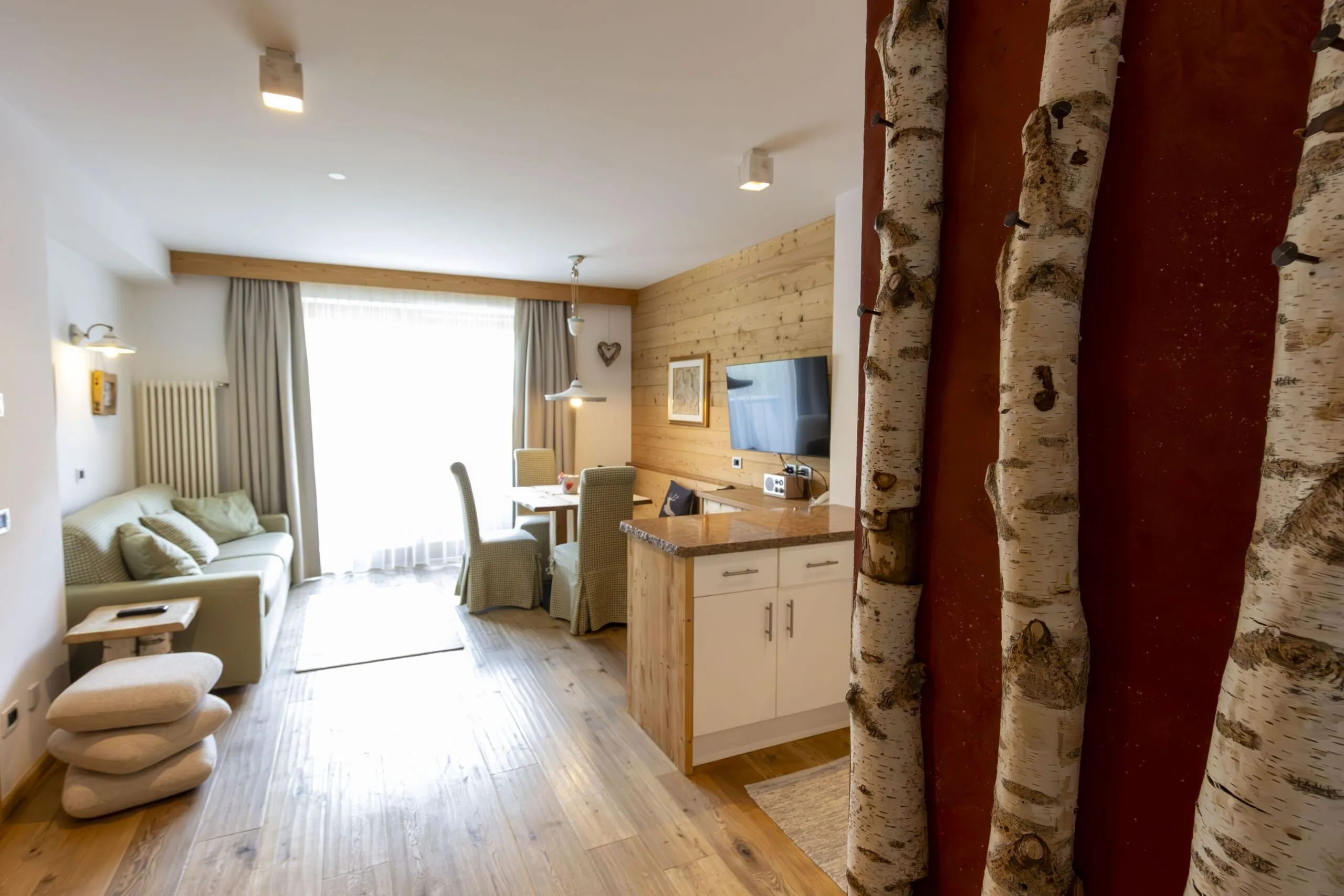 Cozy living area with light wood tones, sofa, dining table, TV on a wood-paneled wall, birch tree decor on the right.