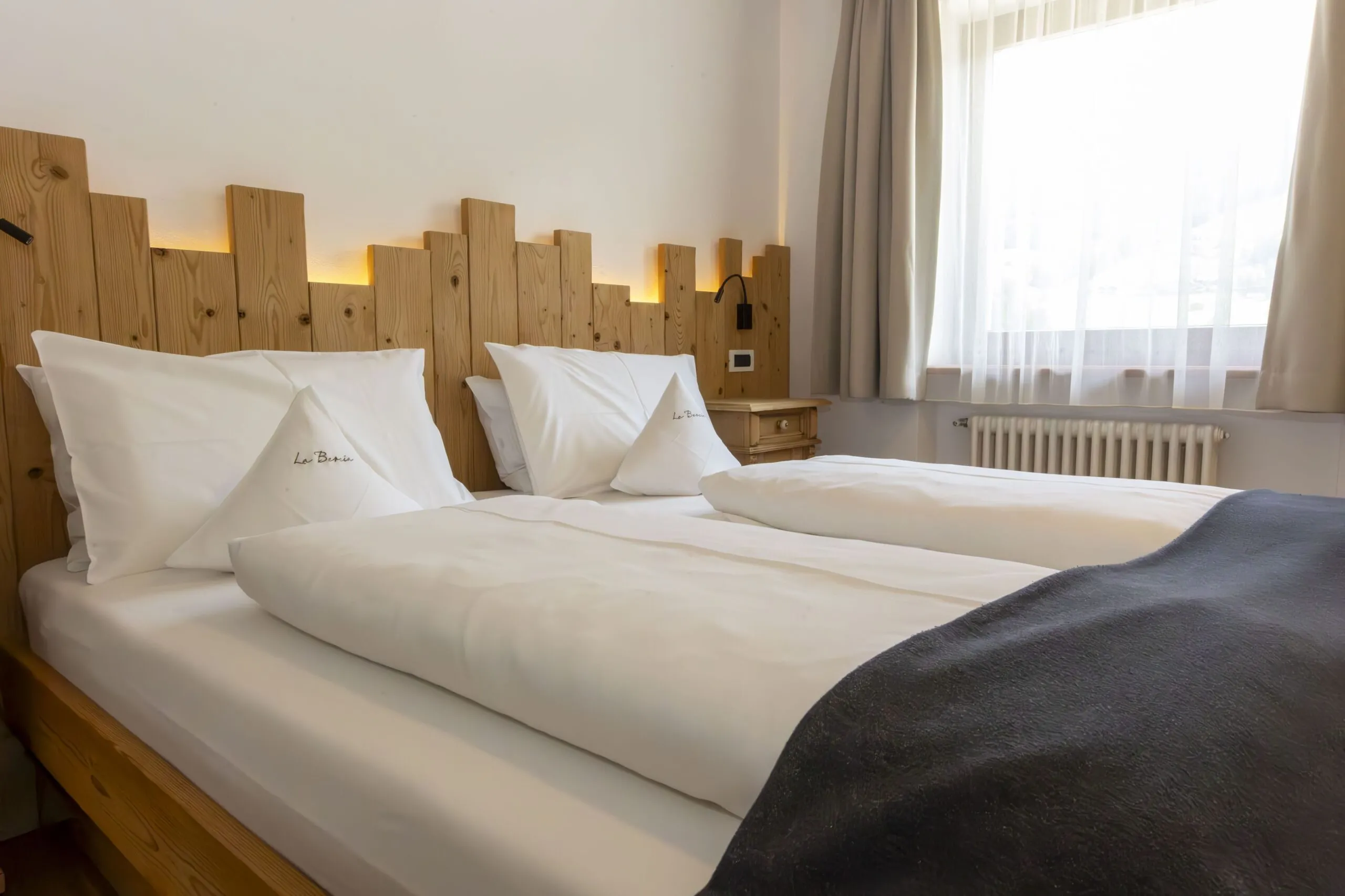 Twin beds with white bedding and wooden headboard, soft lighting, and a large window with sheer curtains.