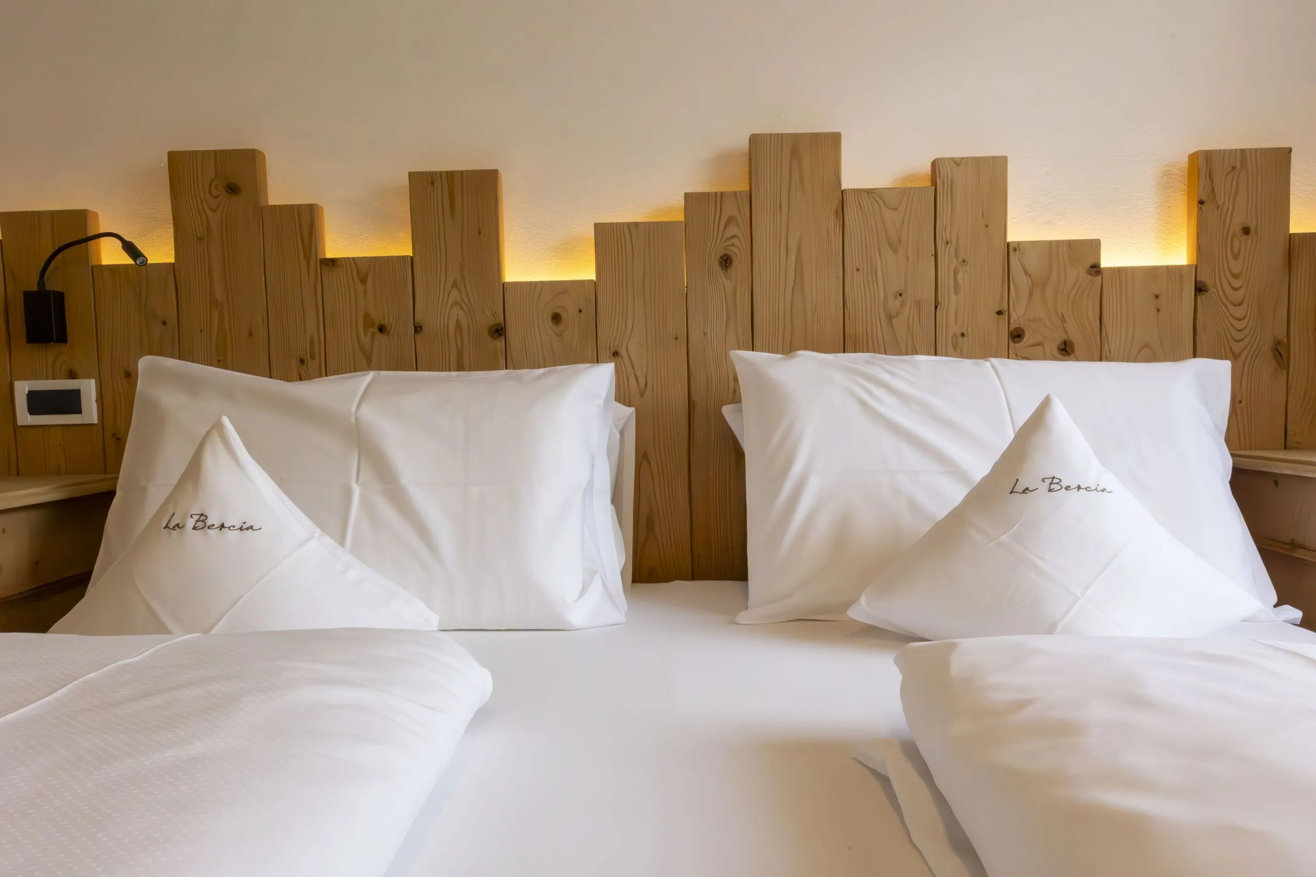 Two white, plush pillows with embroidered “La Berci a” on a wooden headboard and soft warm lighting behind.