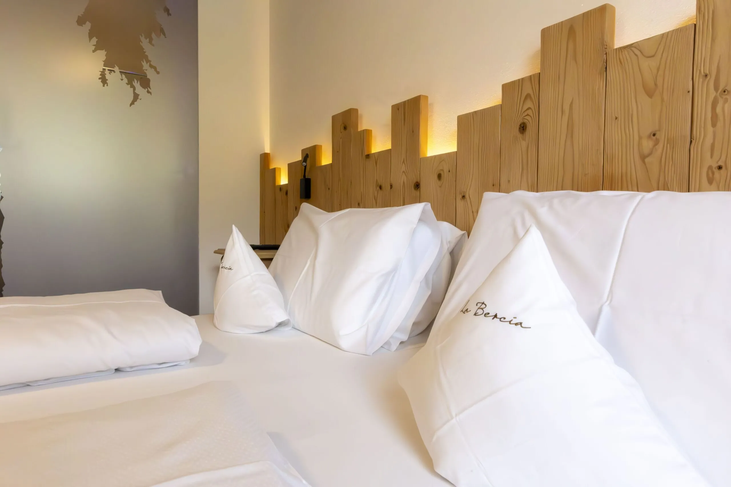 Comfortable hotel bed with white linens and embroidered pillows against a wooden headboard with warm wall lighting.