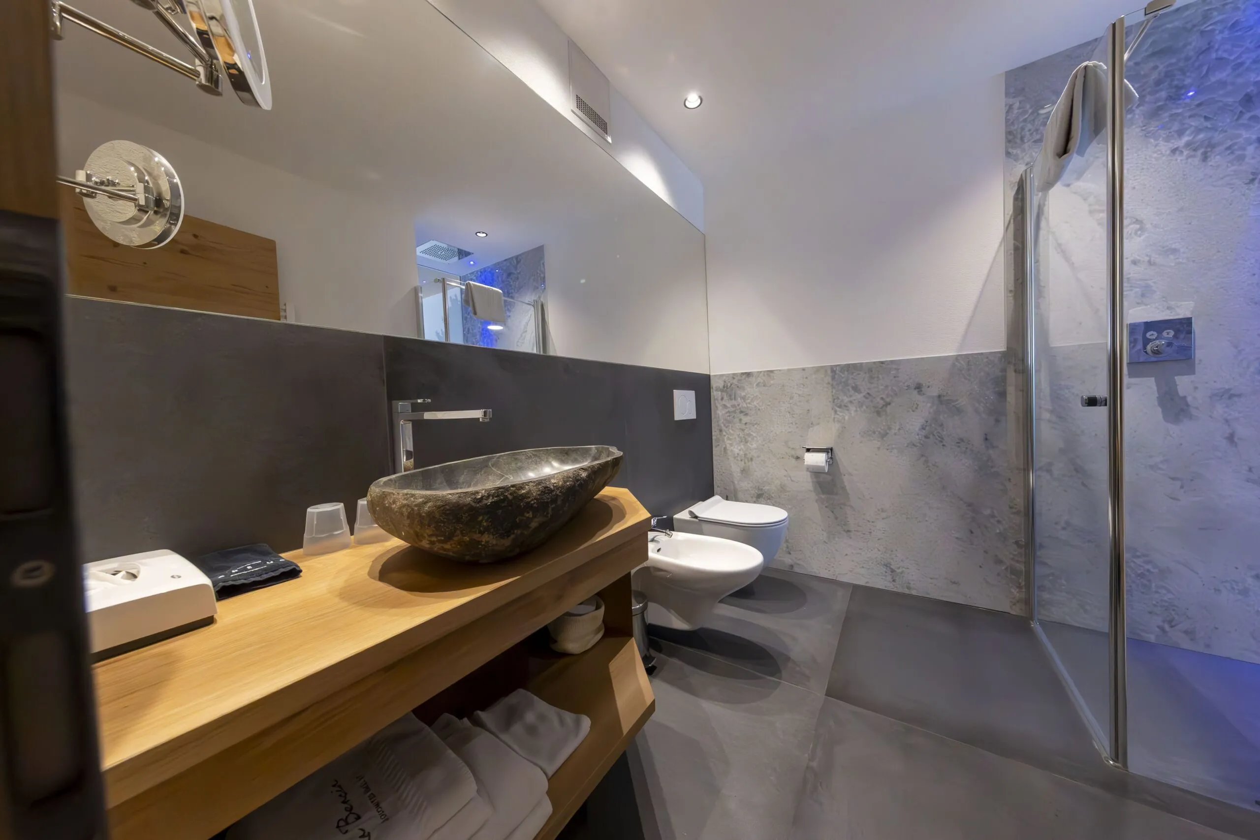 Rustic stone vessel sink on wooden vanity in a modern marble-tiled bathroom with shower.