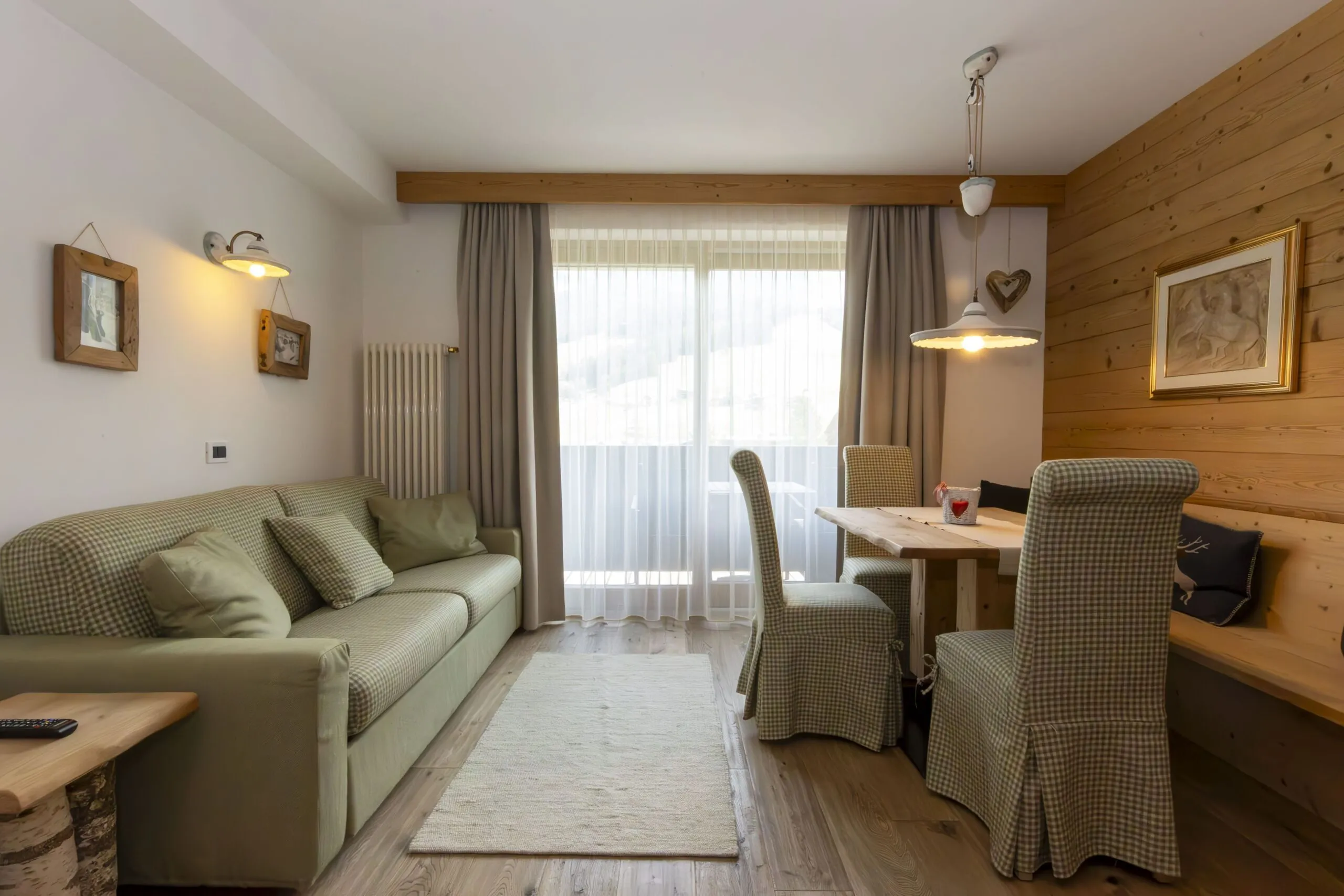 Cozy living and dining area with plaid sofa, wooden walls, and a dining table by a large window.