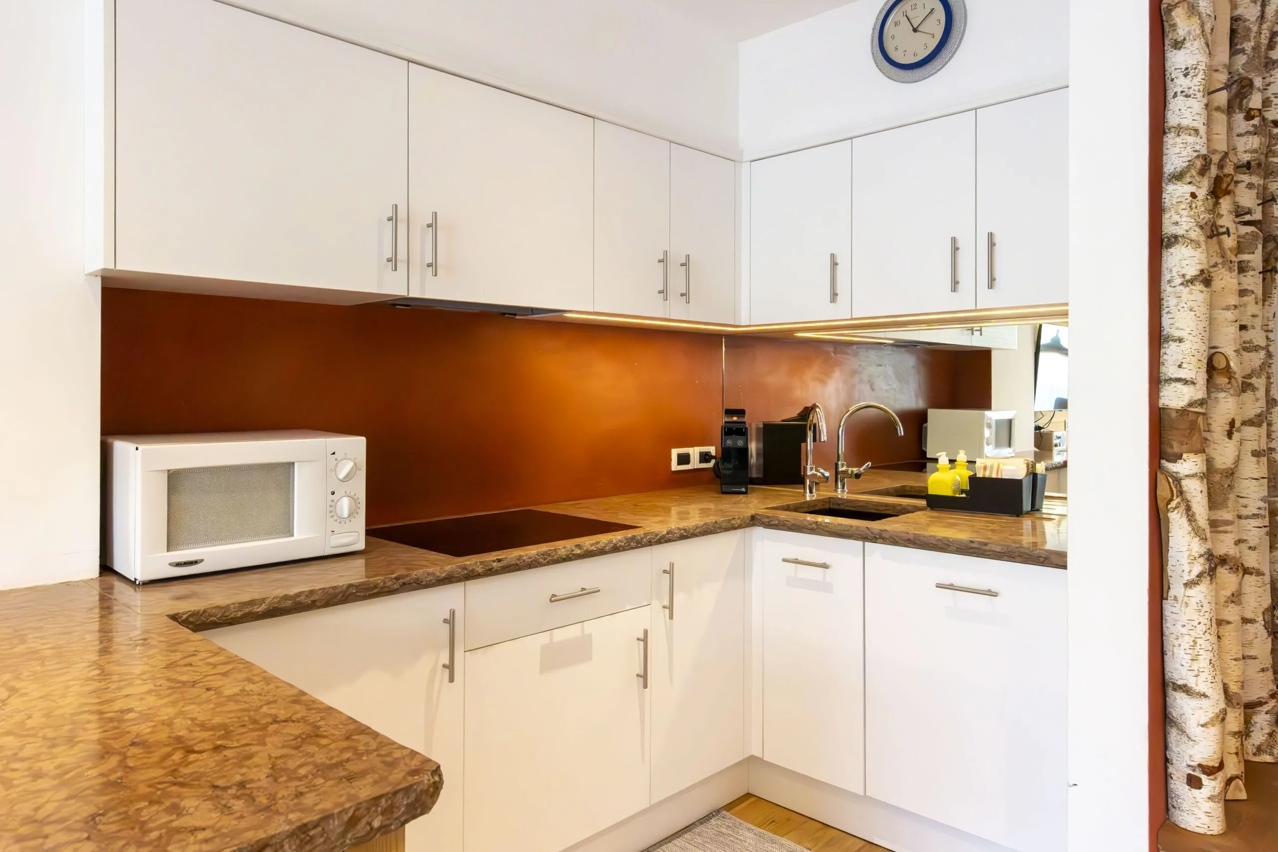 Compact white kitchen with brown marble countertops, upper cabinets, sink, microwave, and coffee station.