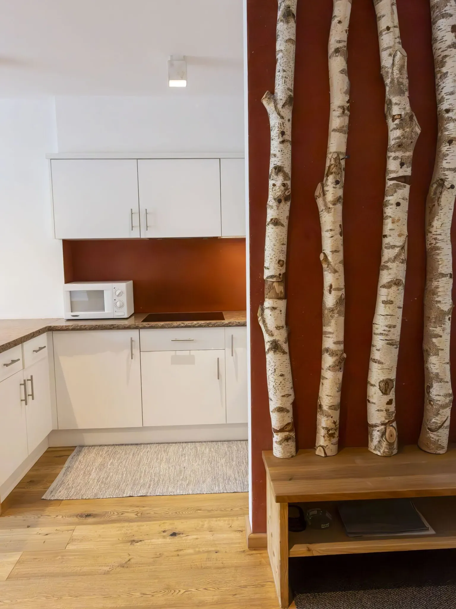 White kitchen with cabinets, microwave, stove, and a wooden bench against a red feature wall with birch tree panels.