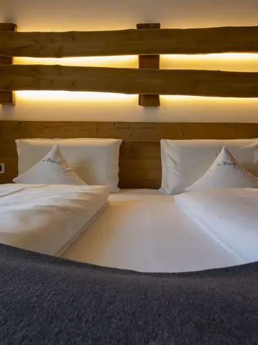 Cozy hotel bed with white linens and decorative pillows, wooden headboard, warm backlit wall, and soft lighting.