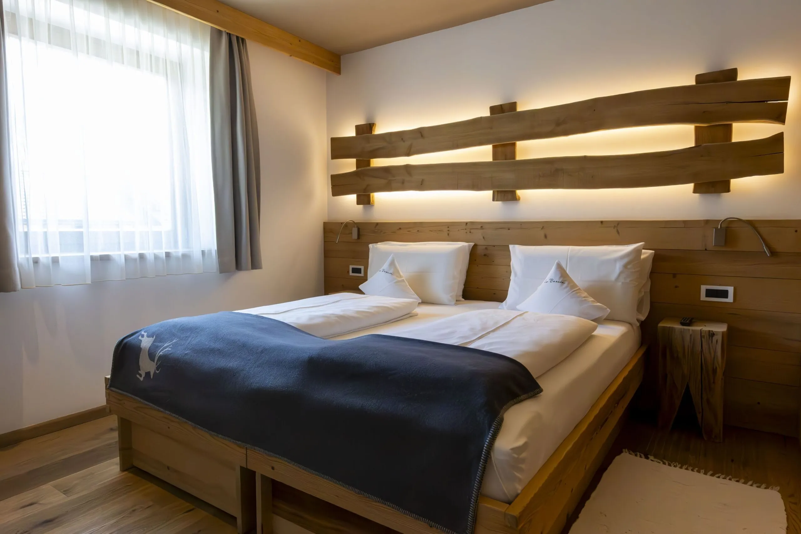 Alpine hotel room with a wooden double bed, split log headboard, warm backlit decor, white bedding, and large window with sheer curtains.