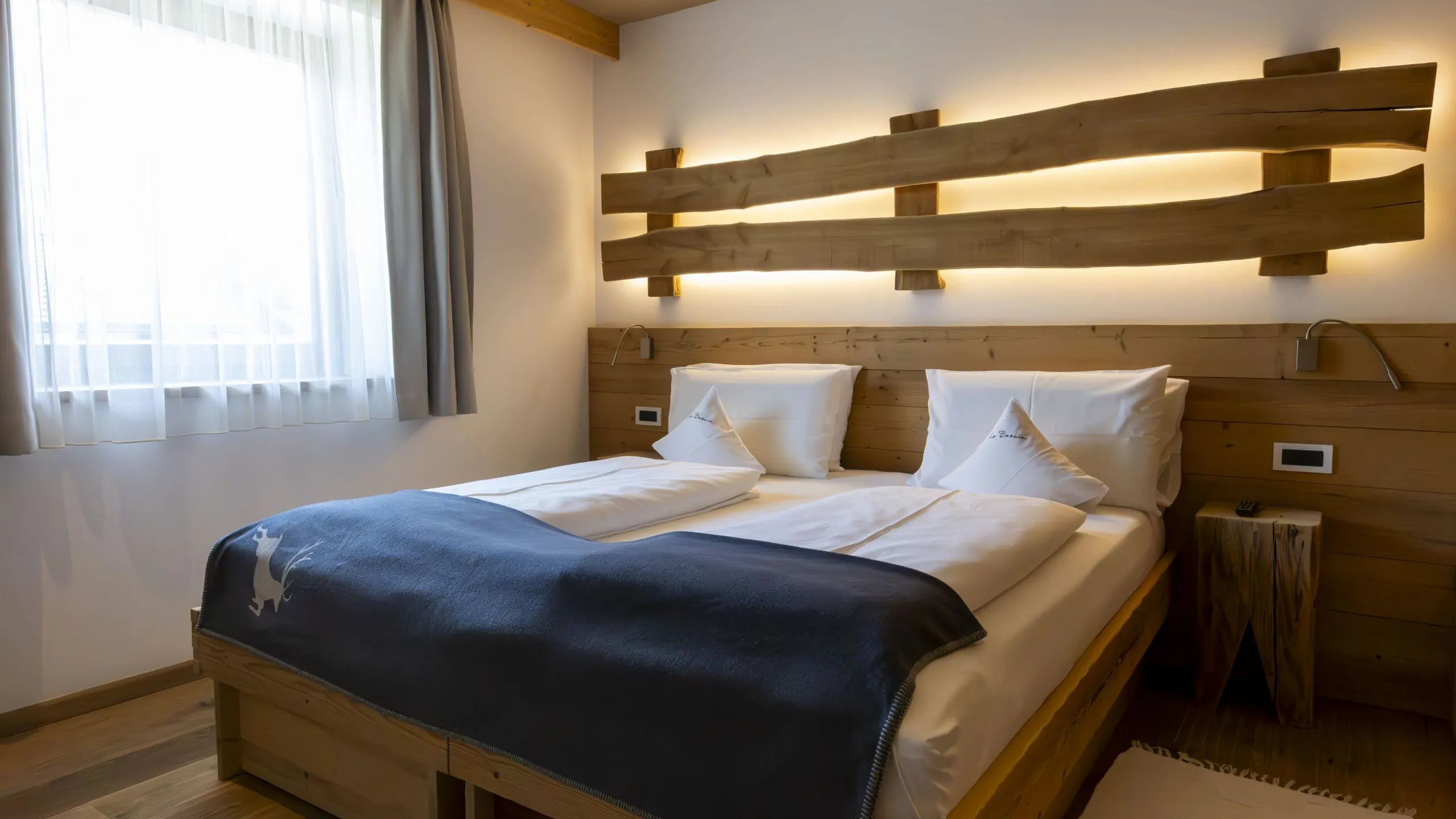 Alpine hotel room with a wooden double bed, split log headboard, warm backlit decor, white bedding, and large window with sheer curtains.