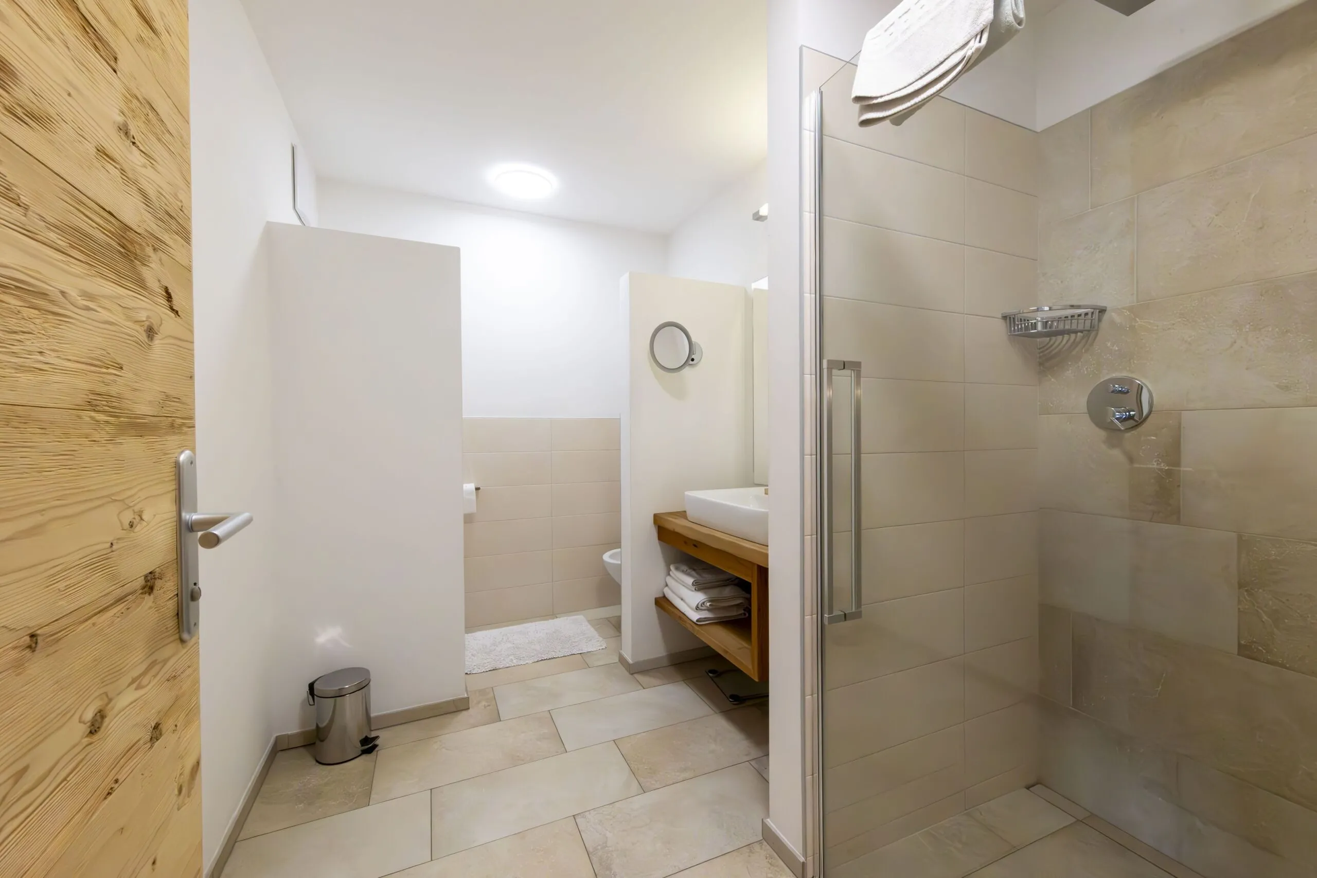 Minimalist bathroom with beige tiled walls, wooden vanity, and glass shower enclosure in a bright, airy space.