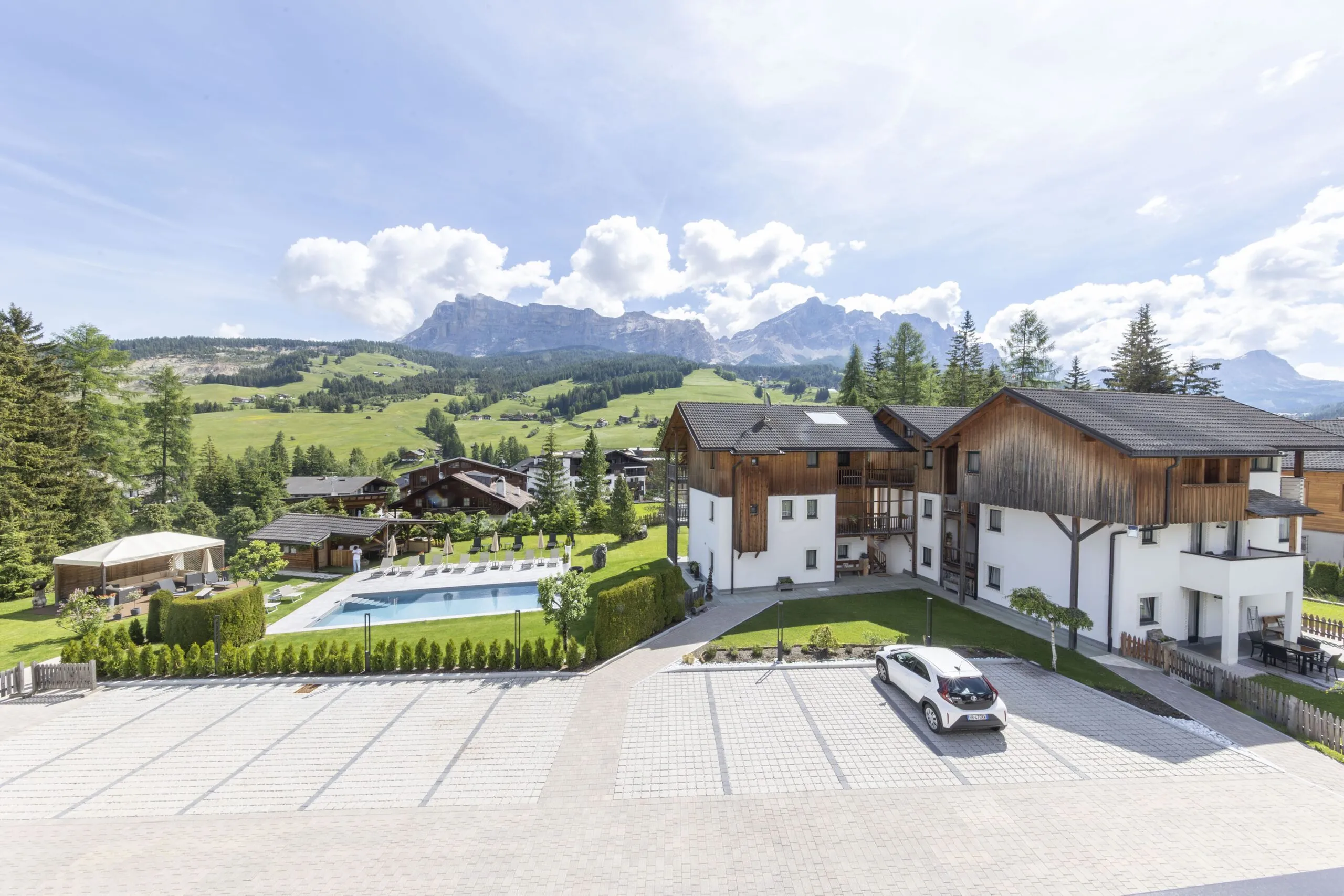 Chalet-style buildings with a pool, green lawn, and mountains in the background under a blue sky.