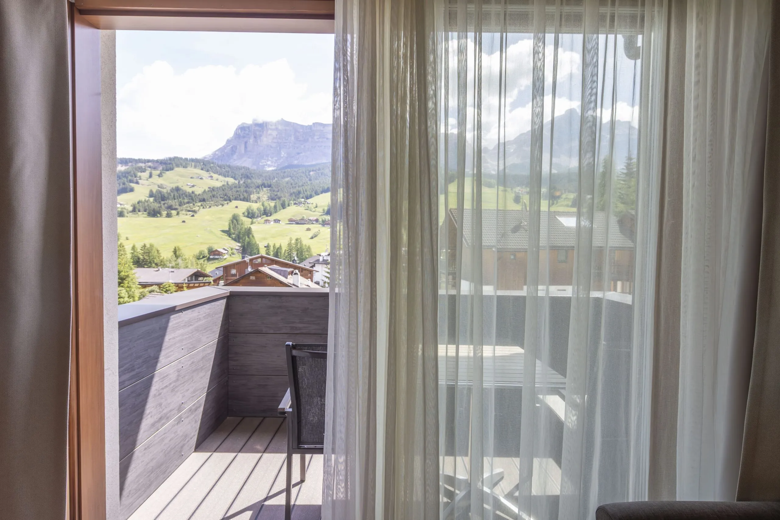 Balcony with wooden railing and chair, view of green valley, hills, and distant mountains through sheer curtains.