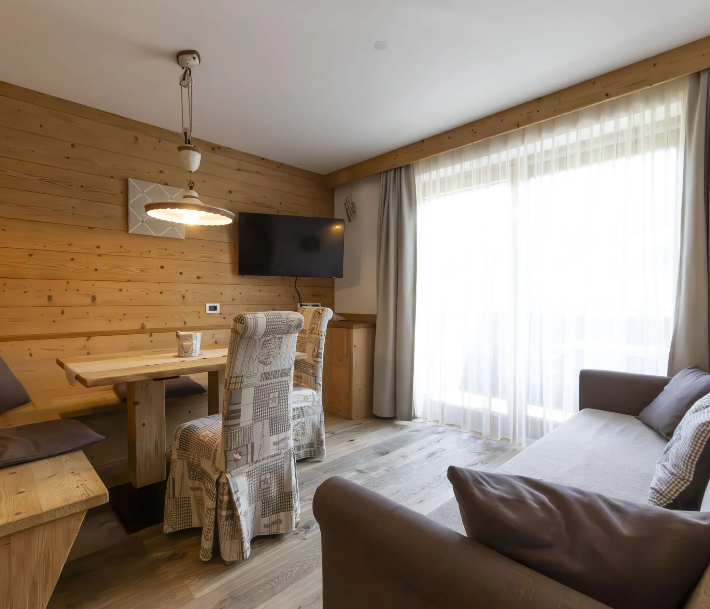 Cozy wood-paneled living/dining area with a bench seat, upholstered chairs, a wall-mounted TV, and large window with sheer curtains.