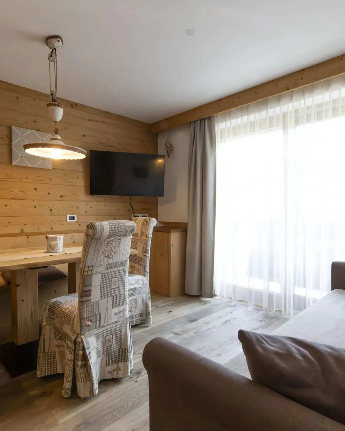 Cozy wood-paneled living/dining area with a bench seat, upholstered chairs, a wall-mounted TV, and large window with sheer curtains.