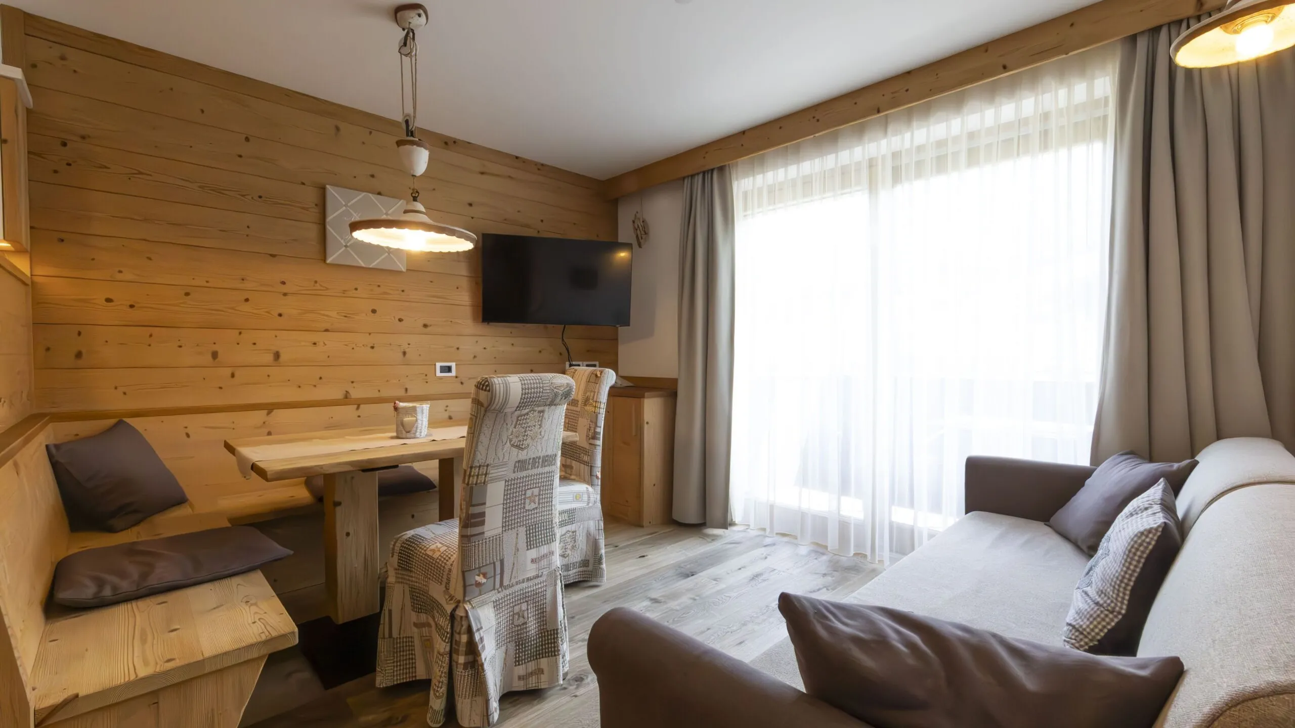 Cozy wood-paneled living/dining area with a bench seat, upholstered chairs, a wall-mounted TV, and large window with sheer curtains.