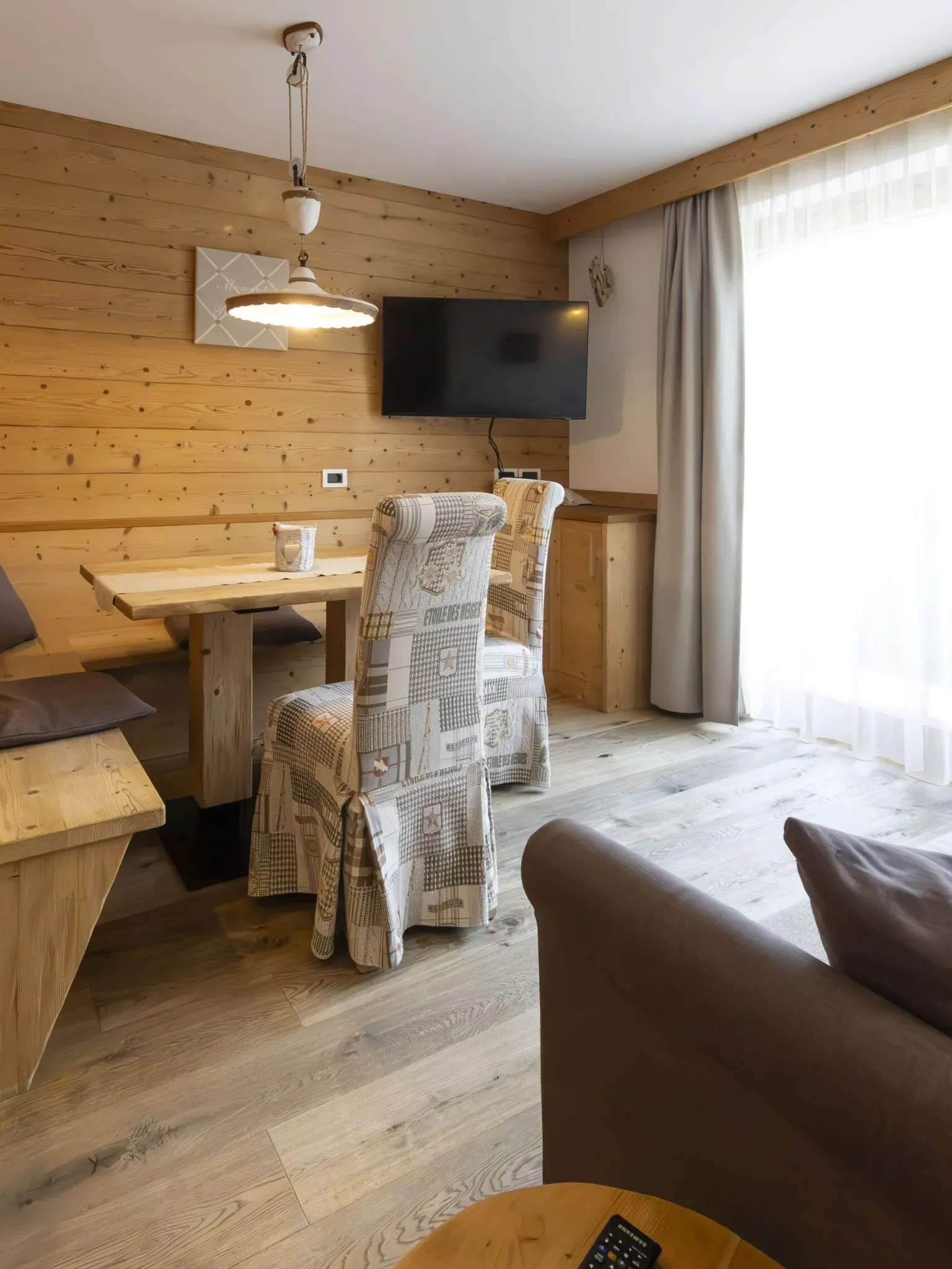 Cozy wooden dining area with a TV on the wall, padded chairs, and a large window with sheer curtains.