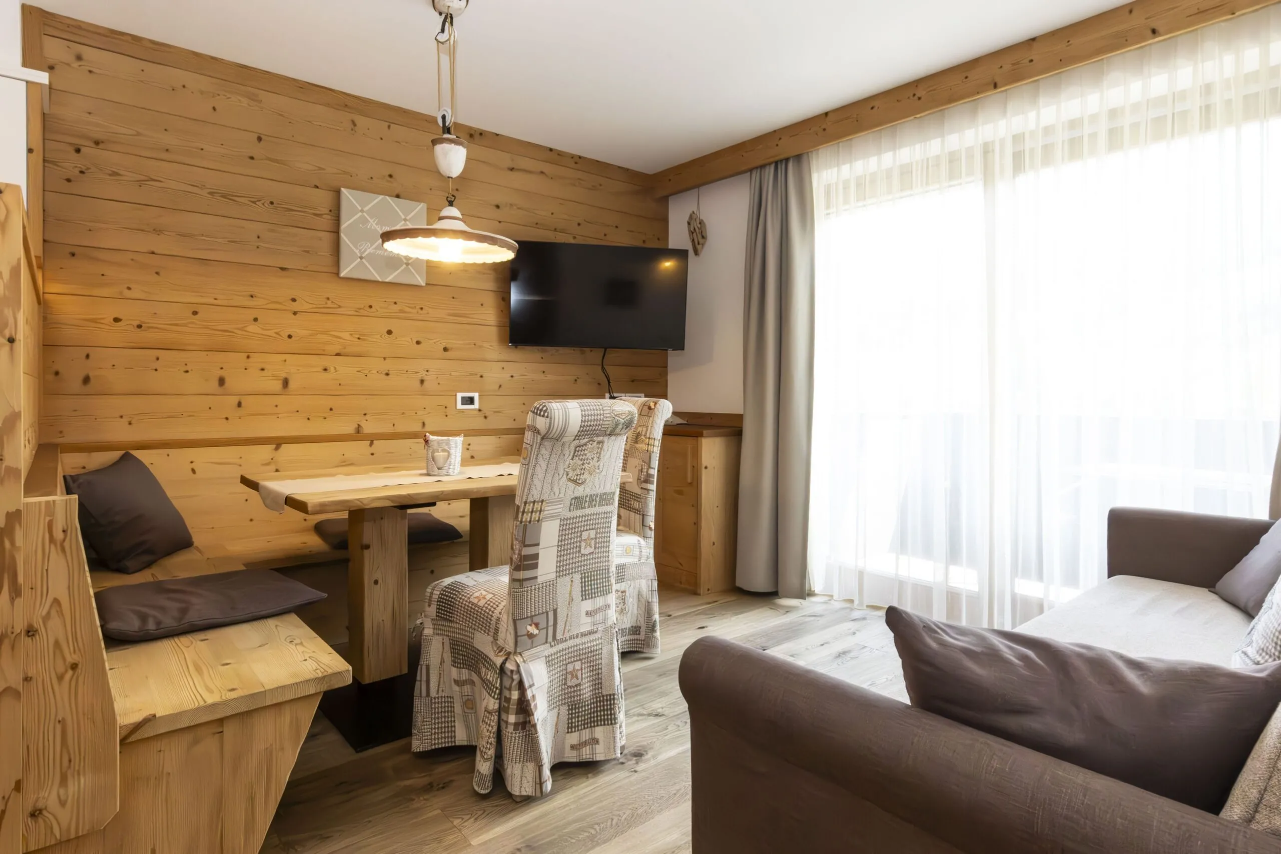 Cozy wooden-lodge living area with a dining nook, wall-mounted TV, and large window with sheer curtains