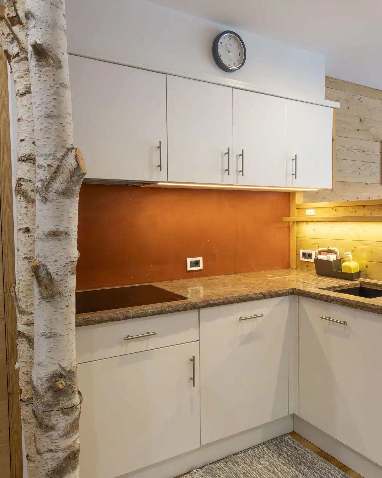 White upper and lower kitchen cabinets with brown backsplash, marble counters, and a birch tree trunk inside the room.