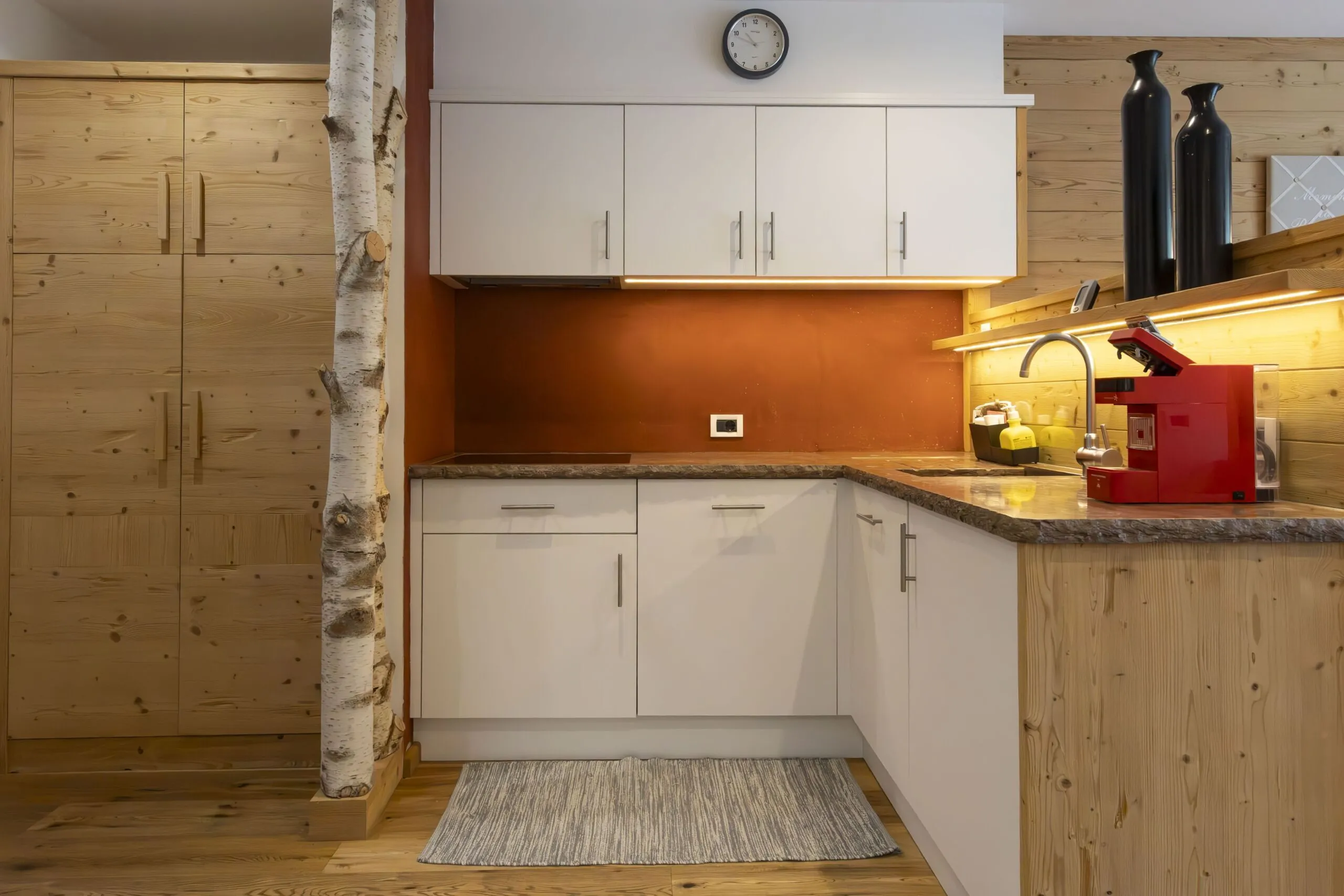 Cozy alpine kitchen with white cabinets, warm wood panels, red espresso machine, and a birch centerpiece.
