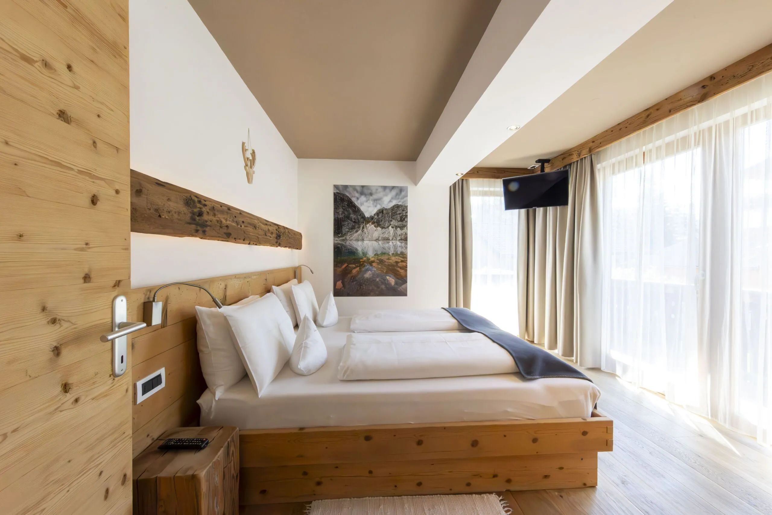 Cozy wooden bedroom with two beds, white linens, large windows, and mountain landscape artwork.