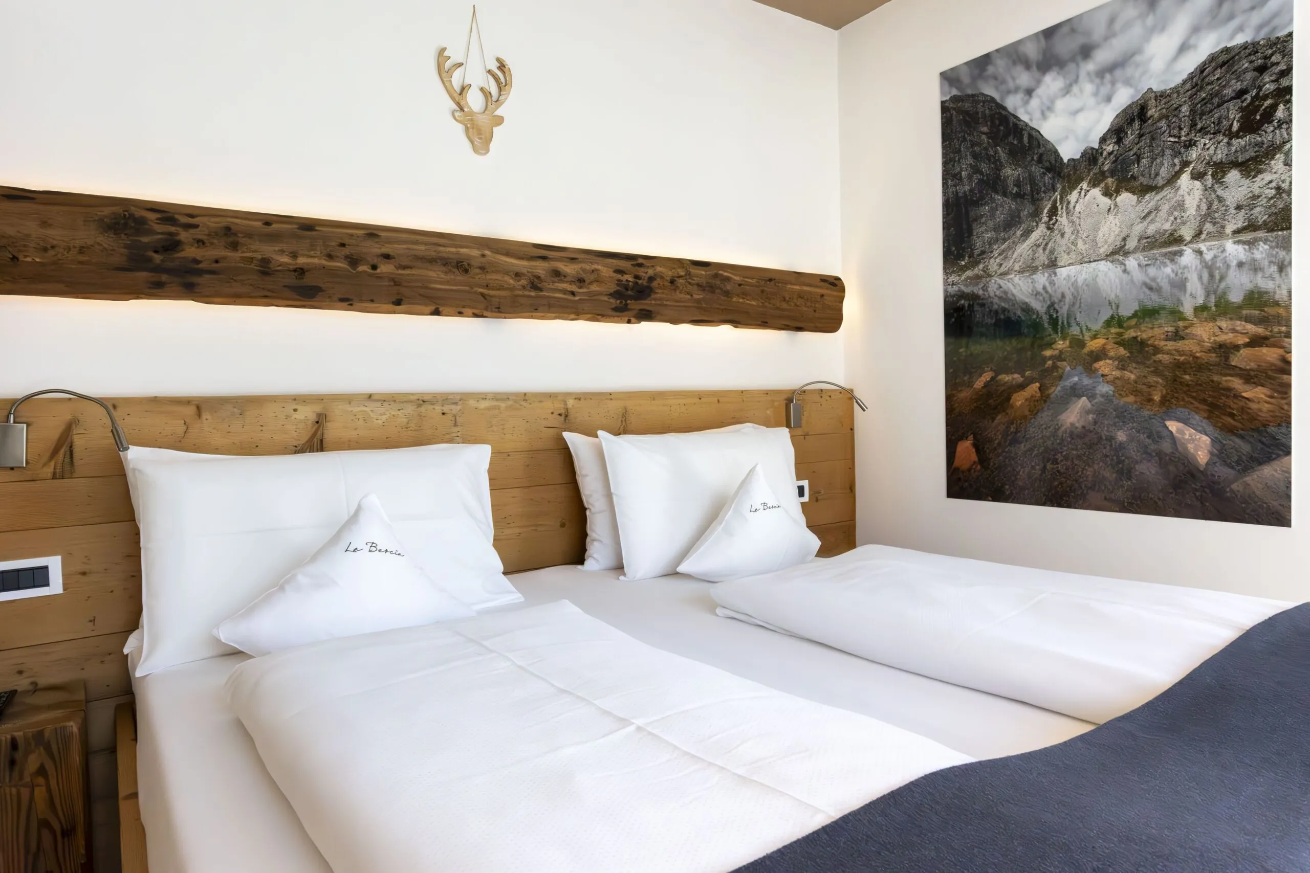 Cozy hotel bedroom with two white beds, wooden headboard, wall lamp, and large mountain photo art.
