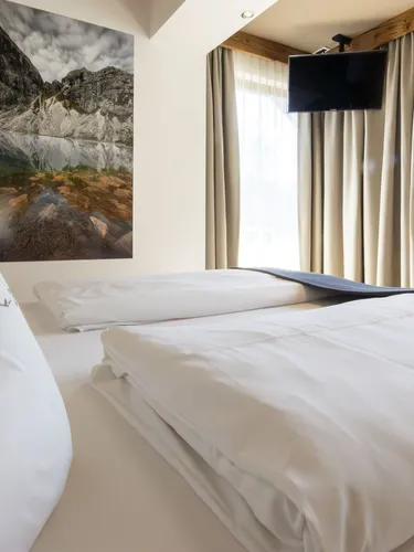 Twin bed with white linens in a bright hotel room, large mountain photo, beige curtains, wall-mounted TV.