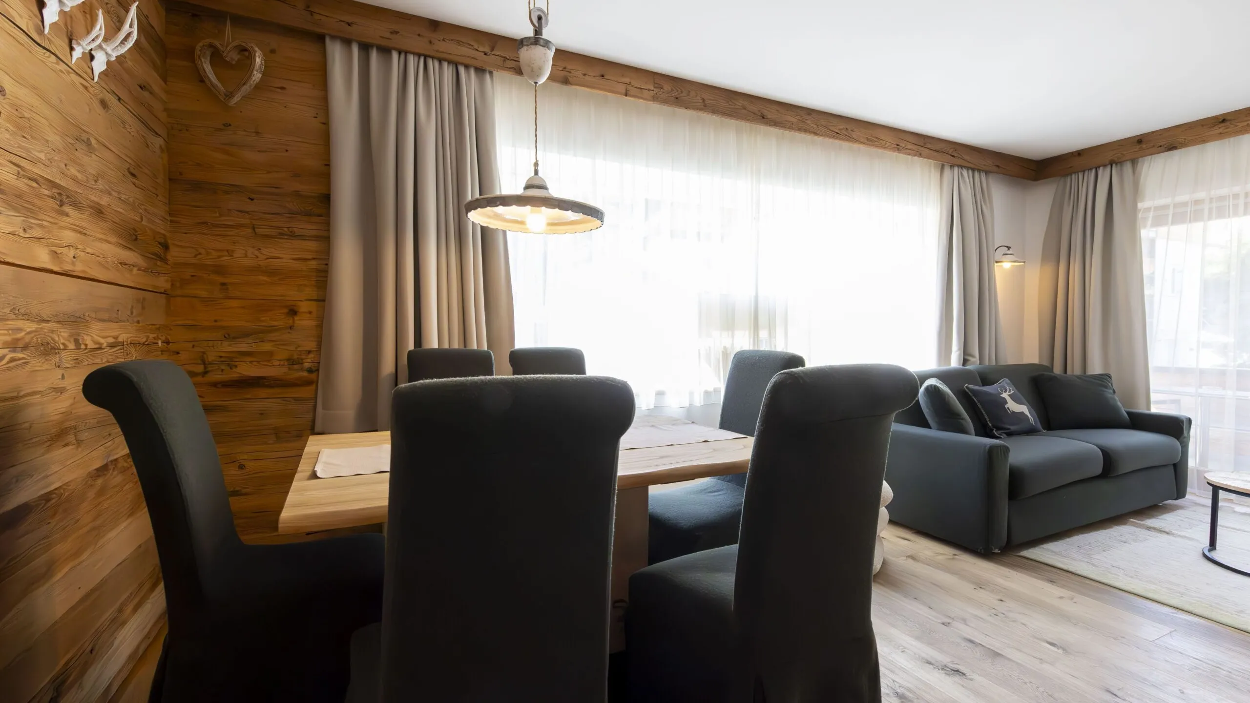 Cozy living and dining area with wood-paneled walls, a wooden dining table and black chairs, plus a gray sofa by large windows with sheer curtains and soft lighting.