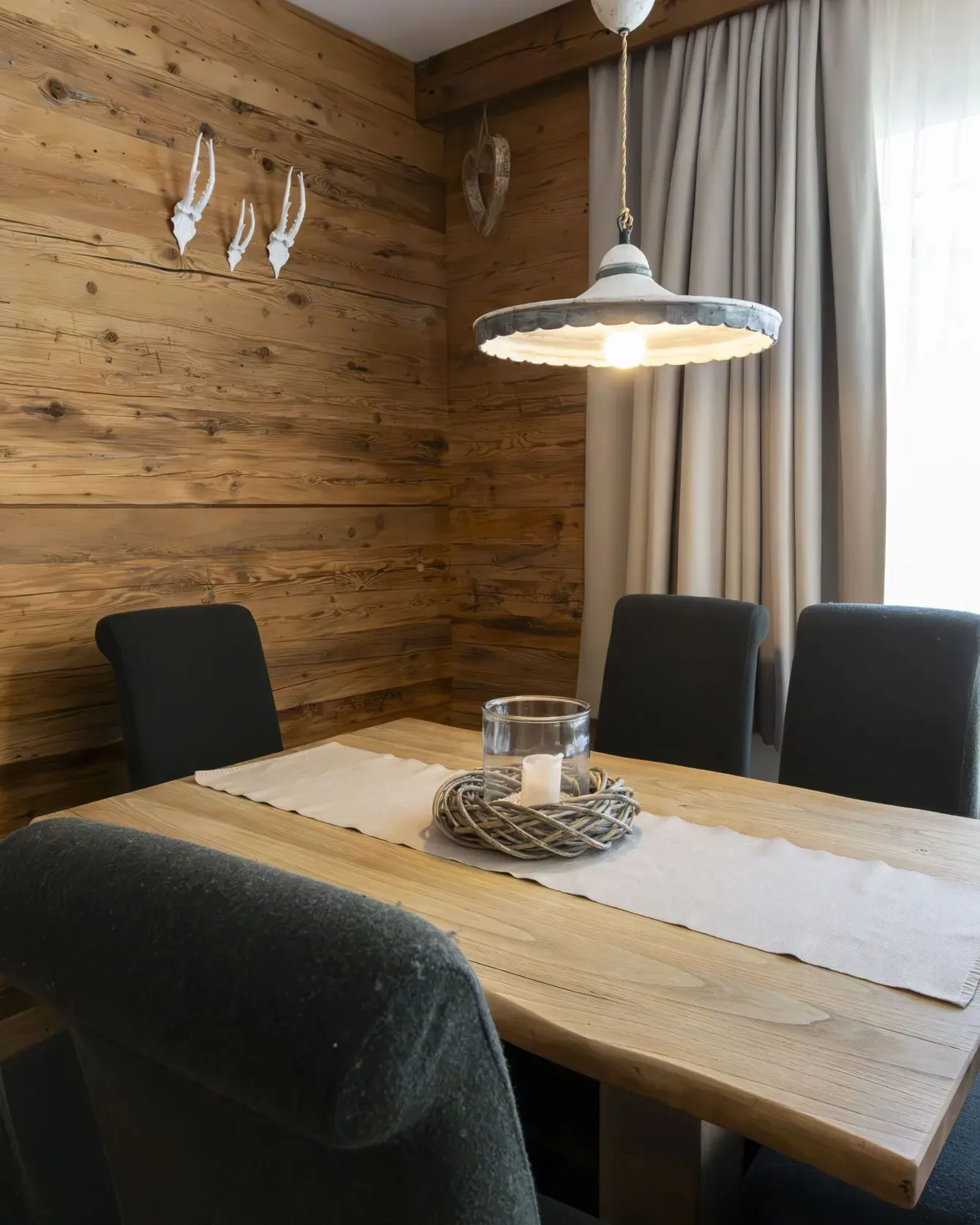 Rustic wooden dining room with a light pendant, beige curtains, and a centerpiece on a wooden table.