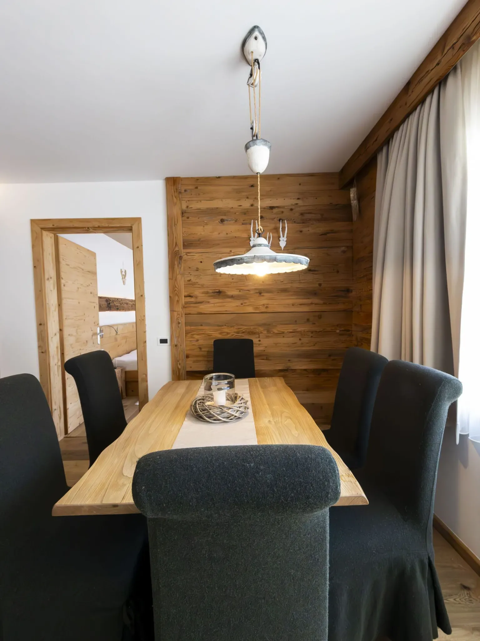 Wooden dining room with a long table, black upholstered chairs, and rustic wood paneled wall under a hanging vintage light fixture.