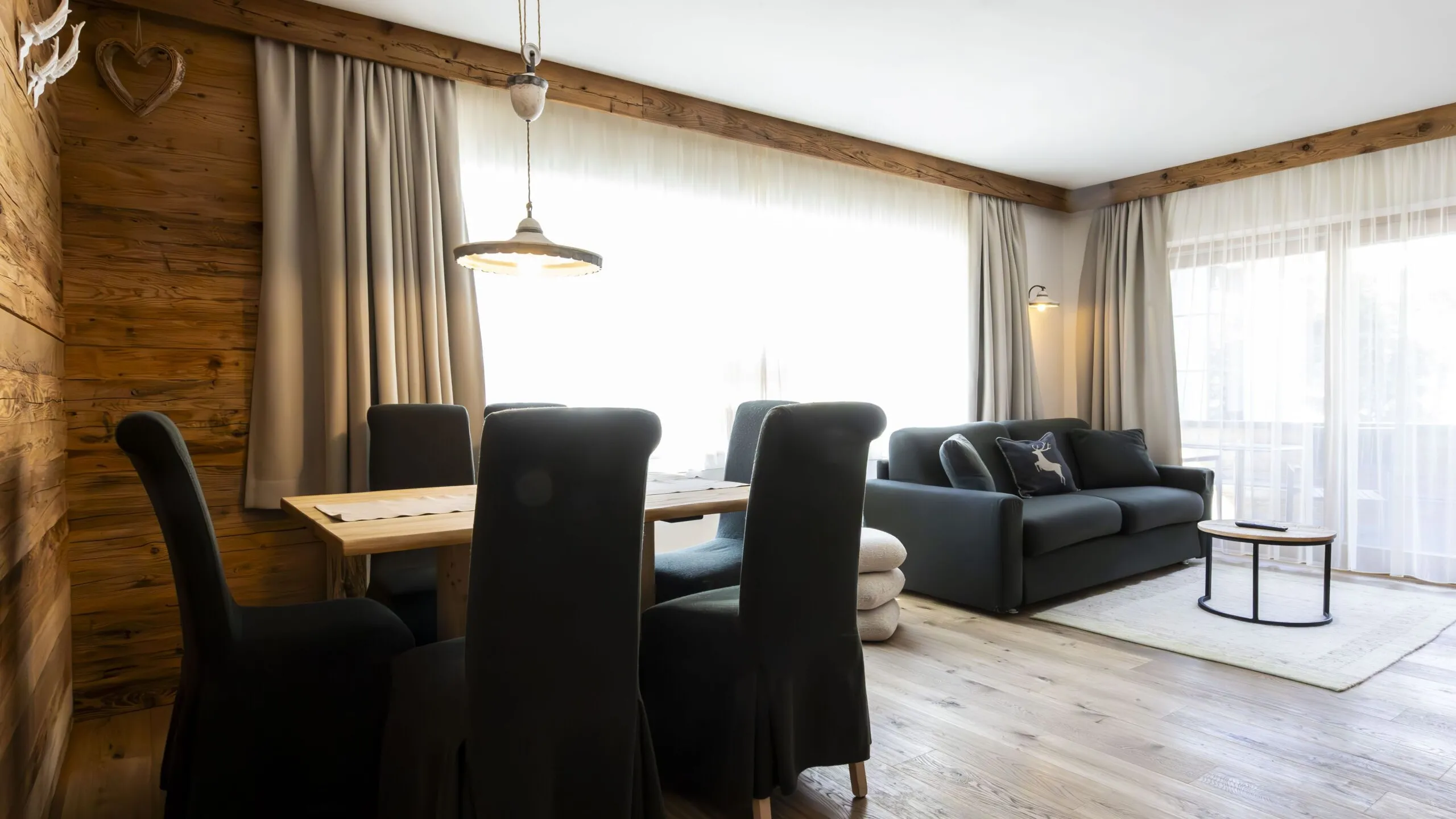 Cozy alpine living and dining area with wooden walls, dark chair covers, and large windows with light curtains.