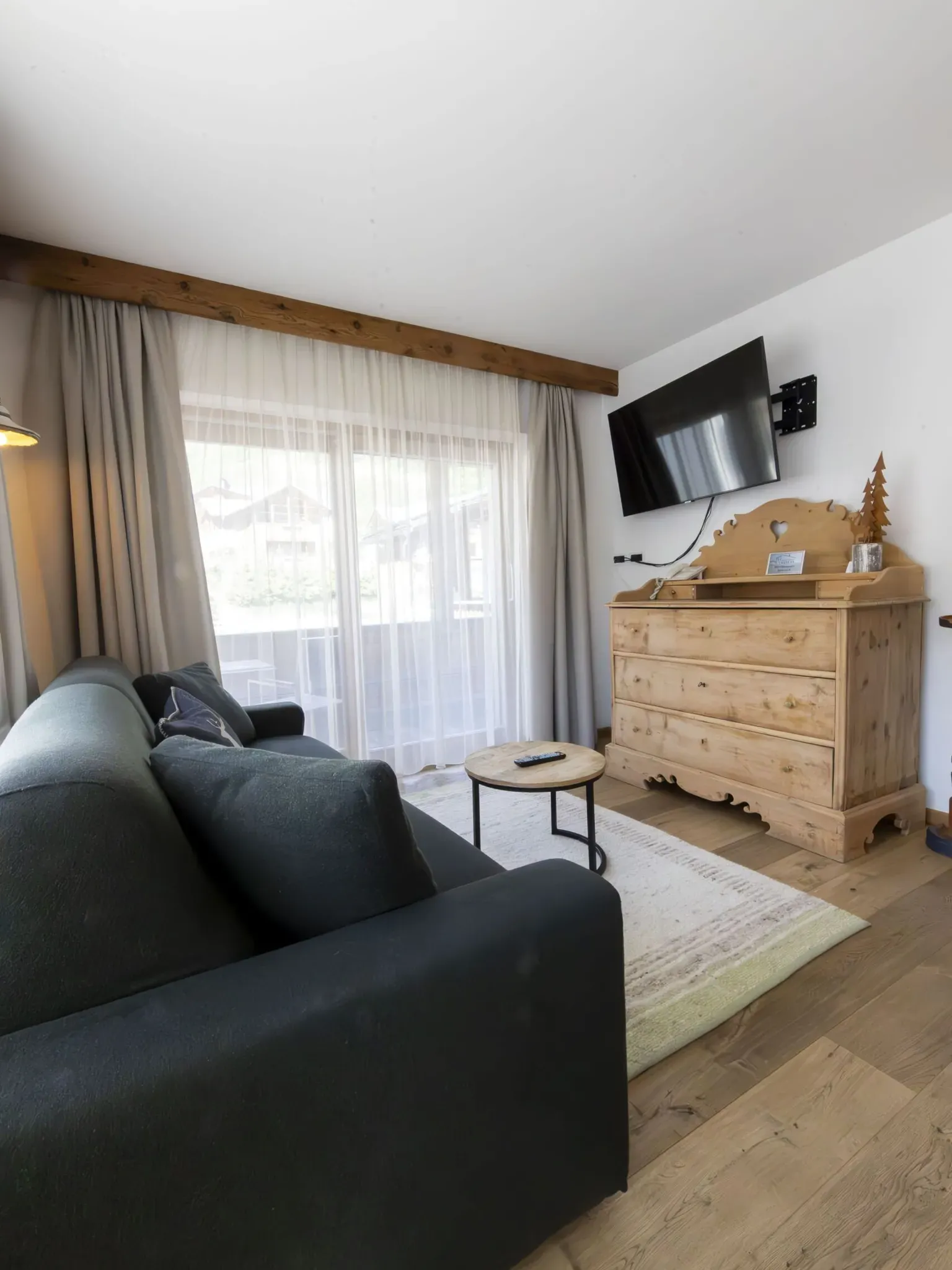 Cozy living room with a dark sofa, wooden dresser, wall-mounted TV, round coffee table, and large glass door with sheer curtains.