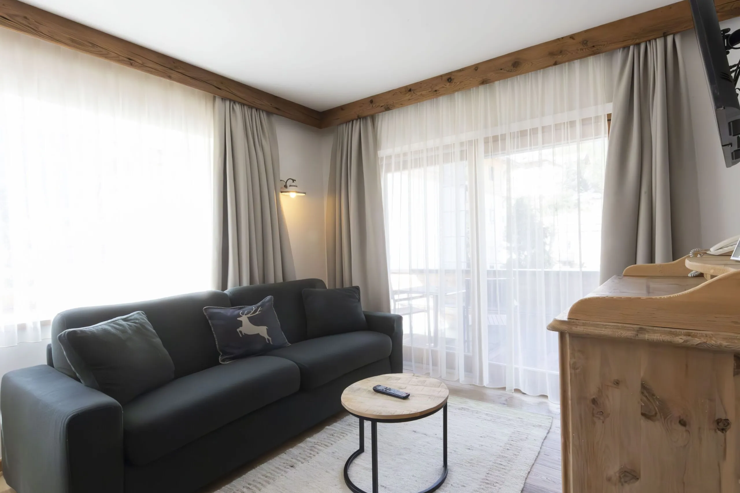 Cozy living room with a dark sofa, decorative pillows, wooden beams, and sheer white curtains by a large window.