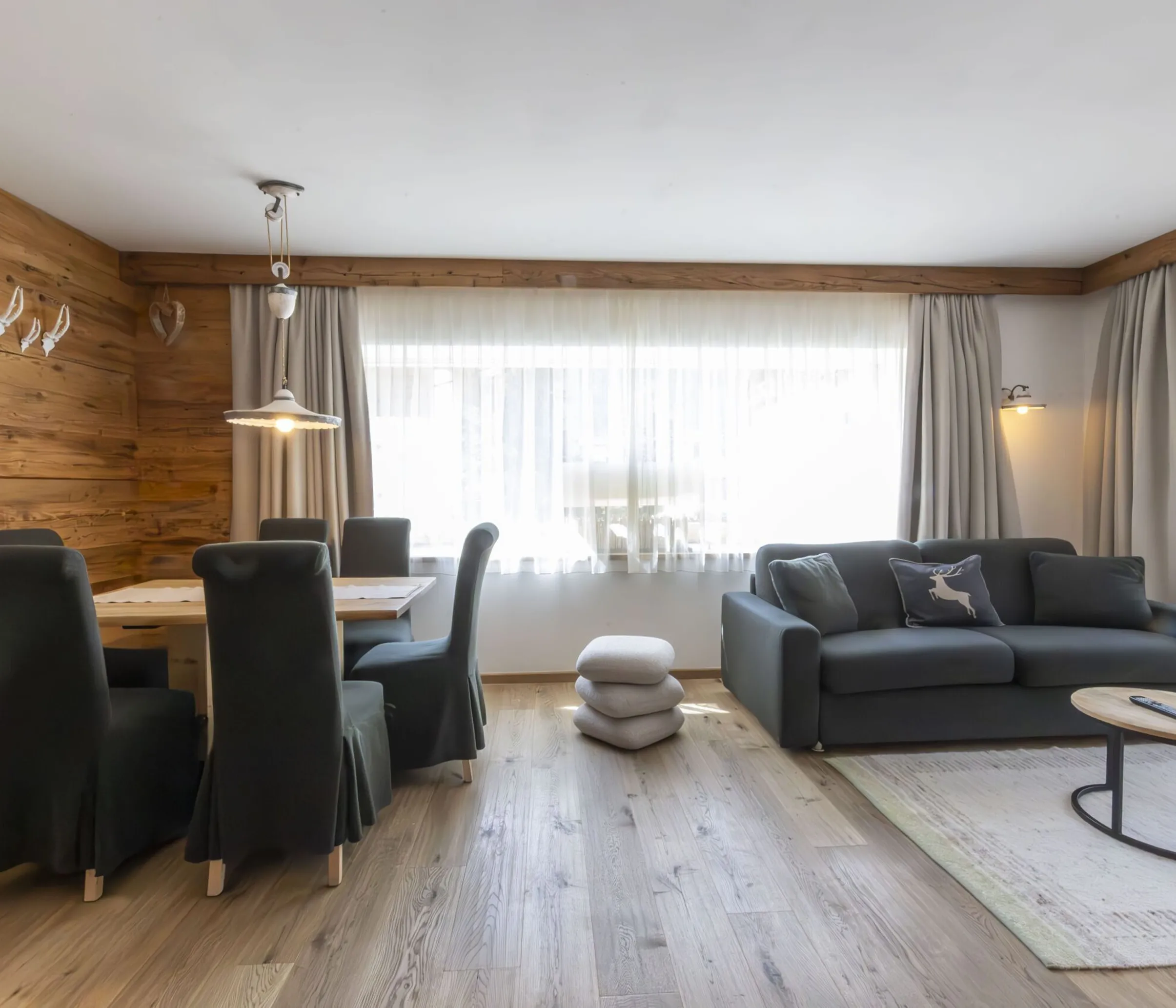 Cozy living and dining area with wood paneled walls, dark upholstered chairs, gray sofa, and large windows with sheer curtains