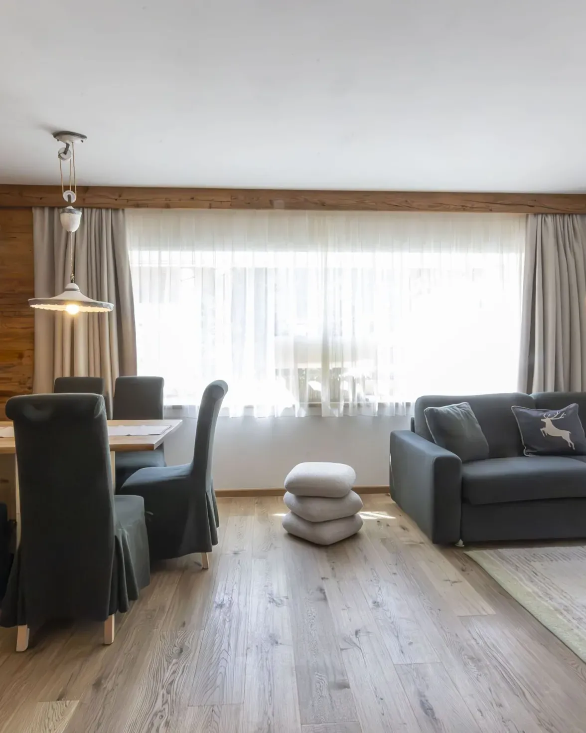Cozy living and dining area with wood paneled walls, dark upholstered chairs, gray sofa, and large windows with sheer curtains