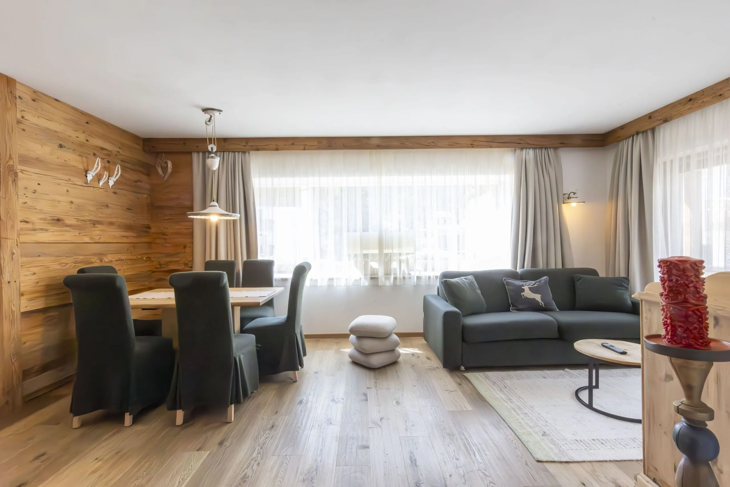 Cozy living and dining area with wooden walls, large windows, gray sofa, and dining table with black chair covers.