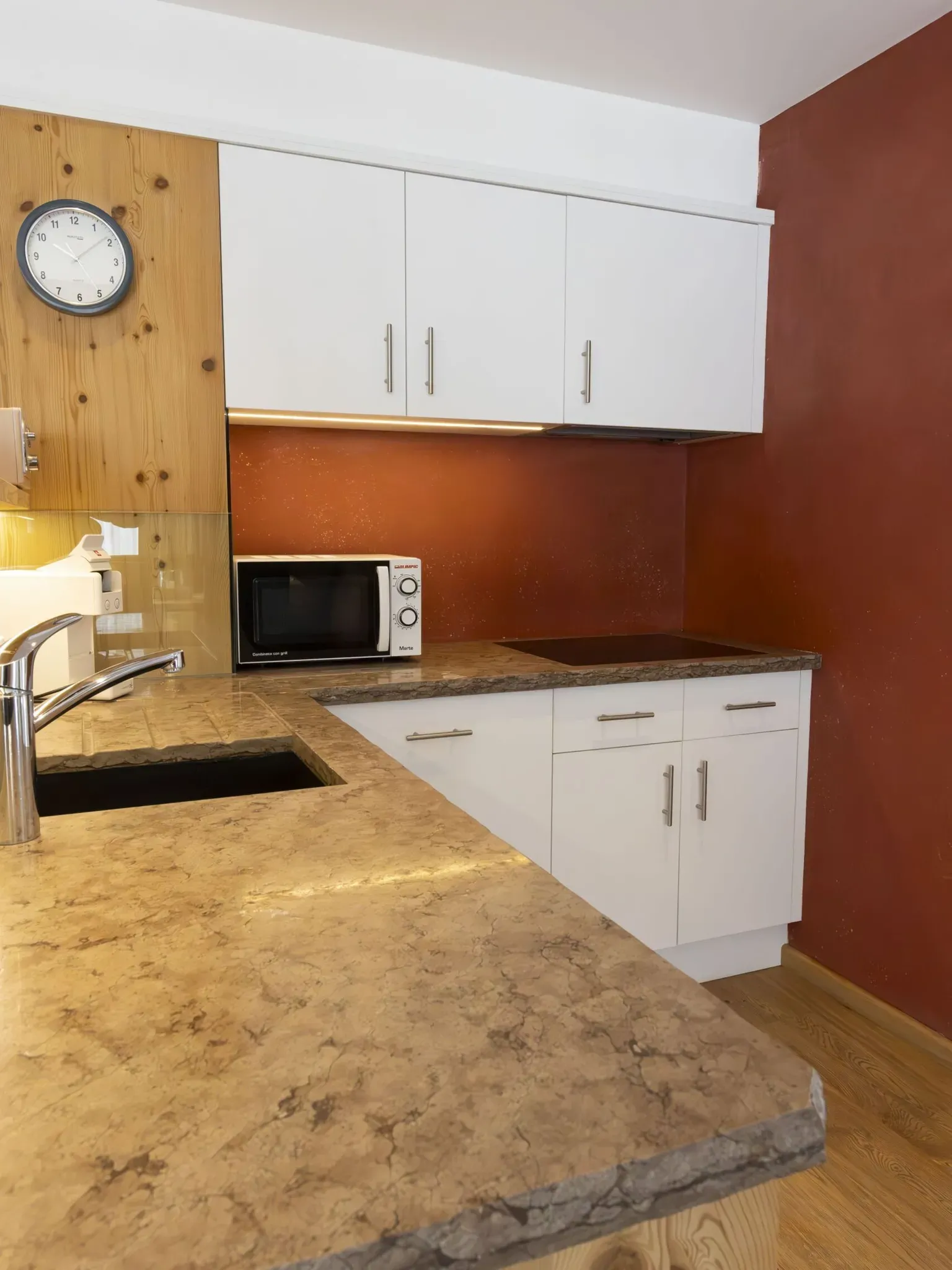 Compact kitchen with white cabinets, marbled brown countertop, microwave, coffee station, and rust-colored wall.