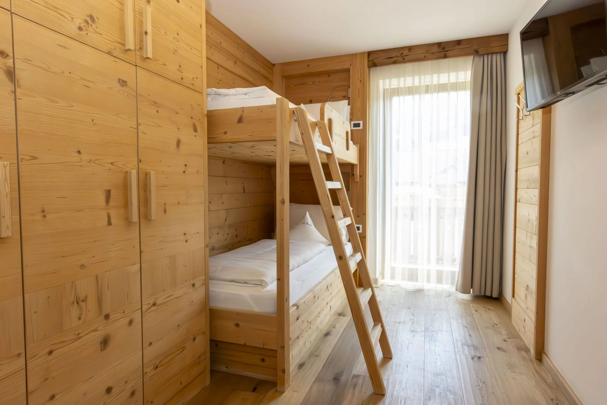 Wooden cabin bedroom with bunk beds, ladder, built-in wardrobes, and large window with sheer curtains.
