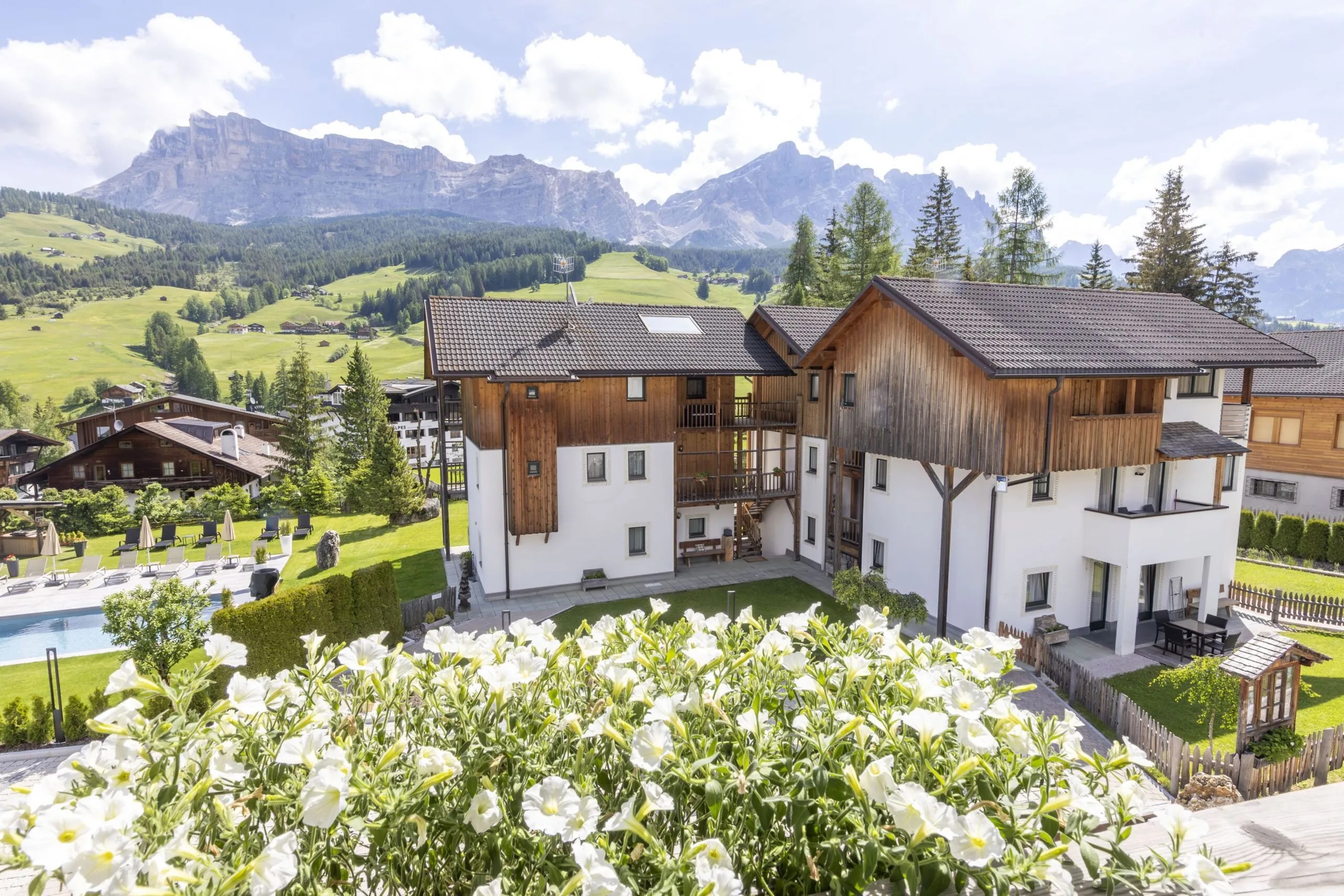 Alpine village hotel with white and wooden facades, surrounding pool, mountains in the background.