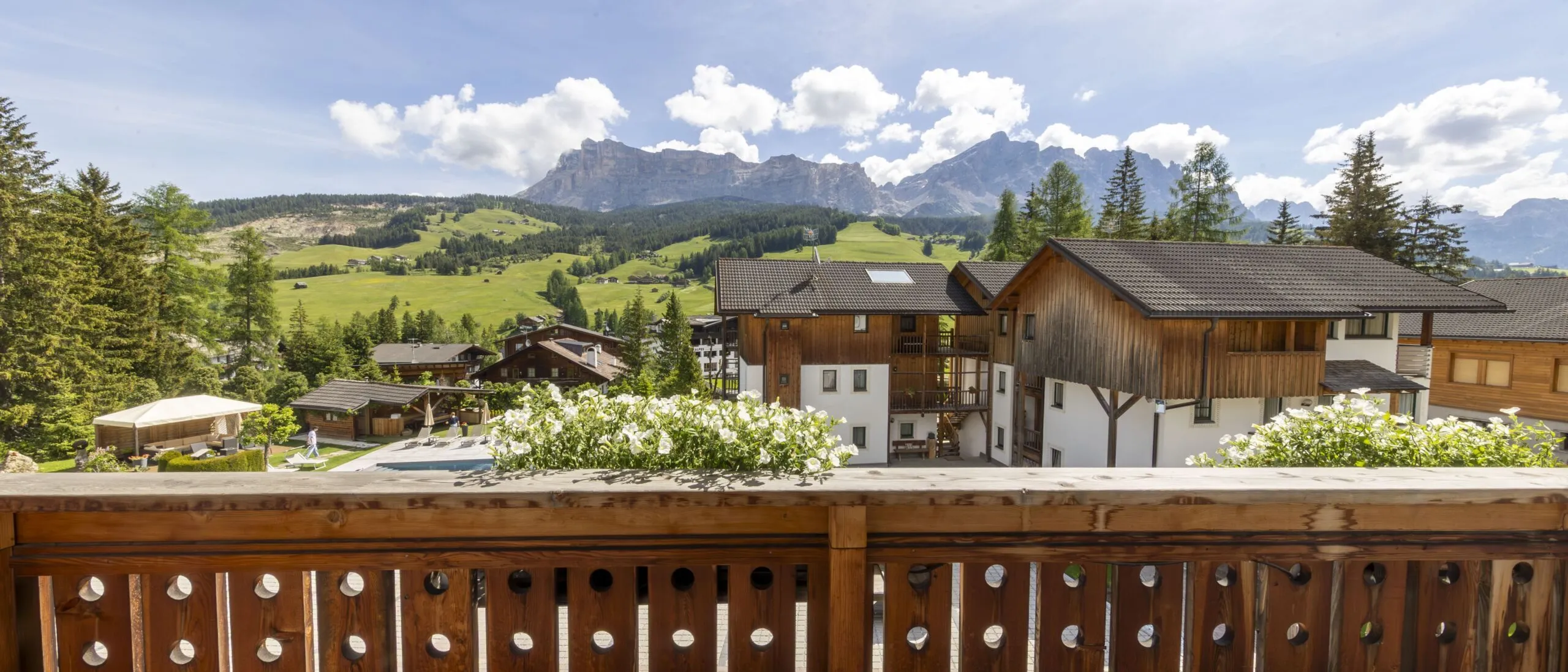 Alpine village view from a wooden balcony with chalet buildings, green fields, and distant mountains.