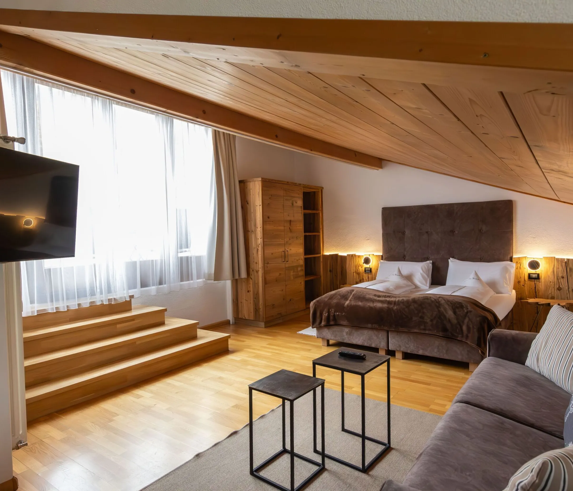 Cozy loft hotel room with wooden ceiling, large bed, storage cabinet, and sofa near a window with sheer curtains