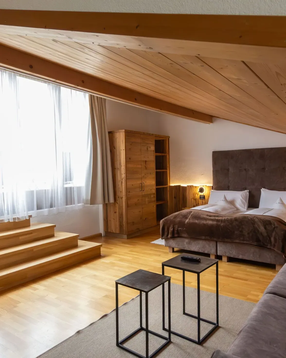 Cozy loft hotel room with wooden ceiling, large bed, storage cabinet, and sofa near a window with sheer curtains