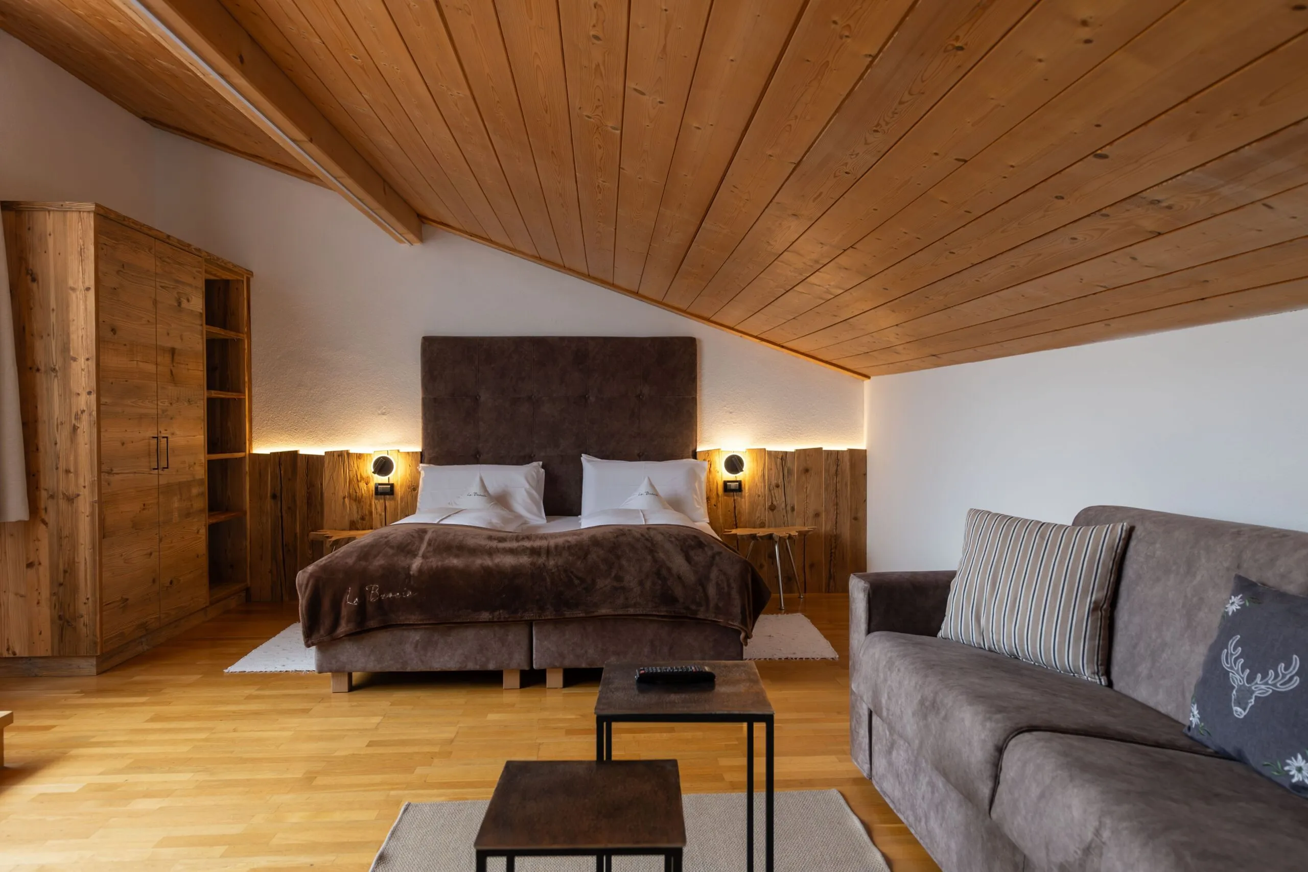 Cozy alpine bedroom with wooden ceiling, large bed, brown headboard, nightstands, wardrobe, and a gray sofa area.