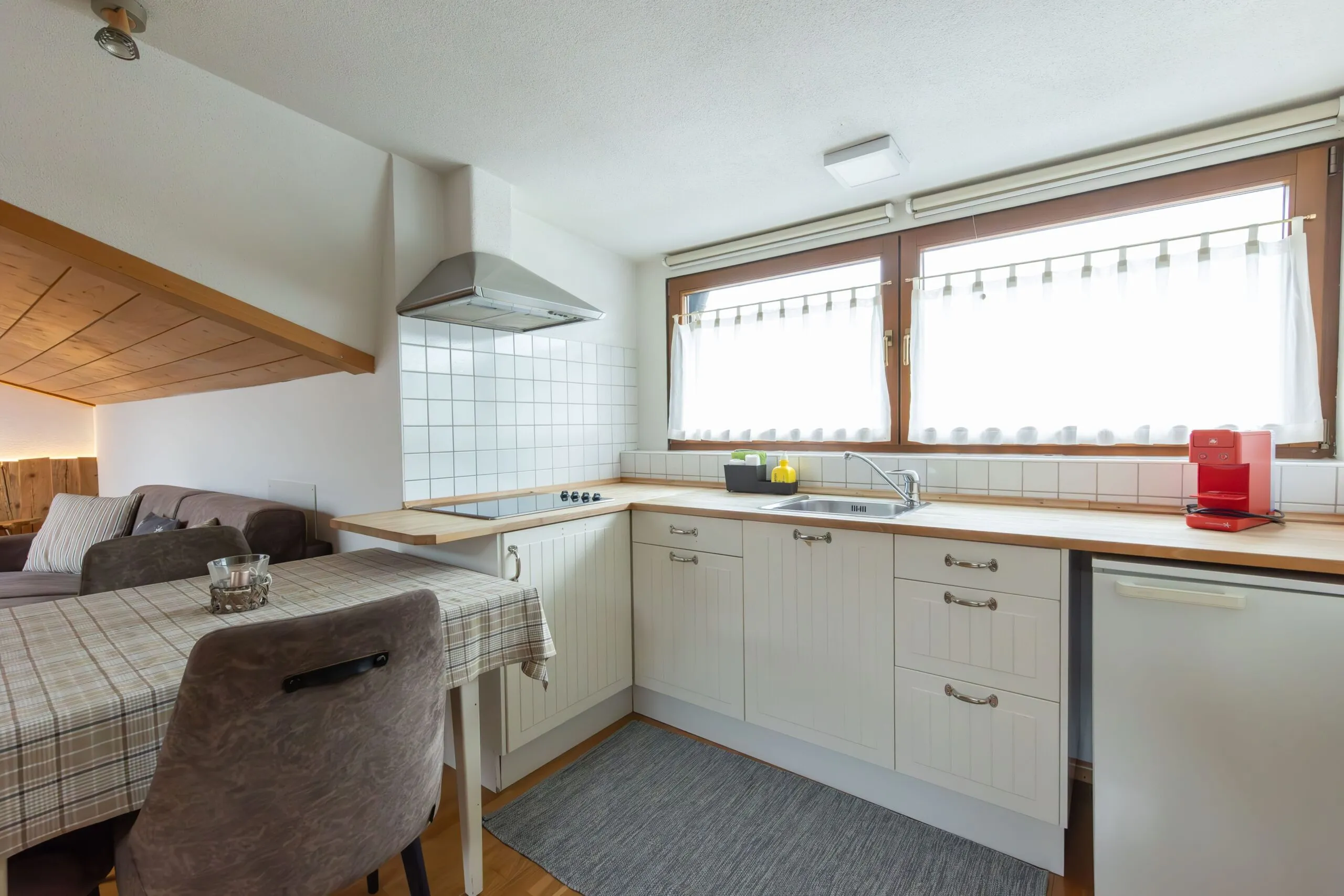 Bright modern L-shaped kitchen with white cabinets, tiled backsplash, wooden countertop, sink, and large windows with white curtains.