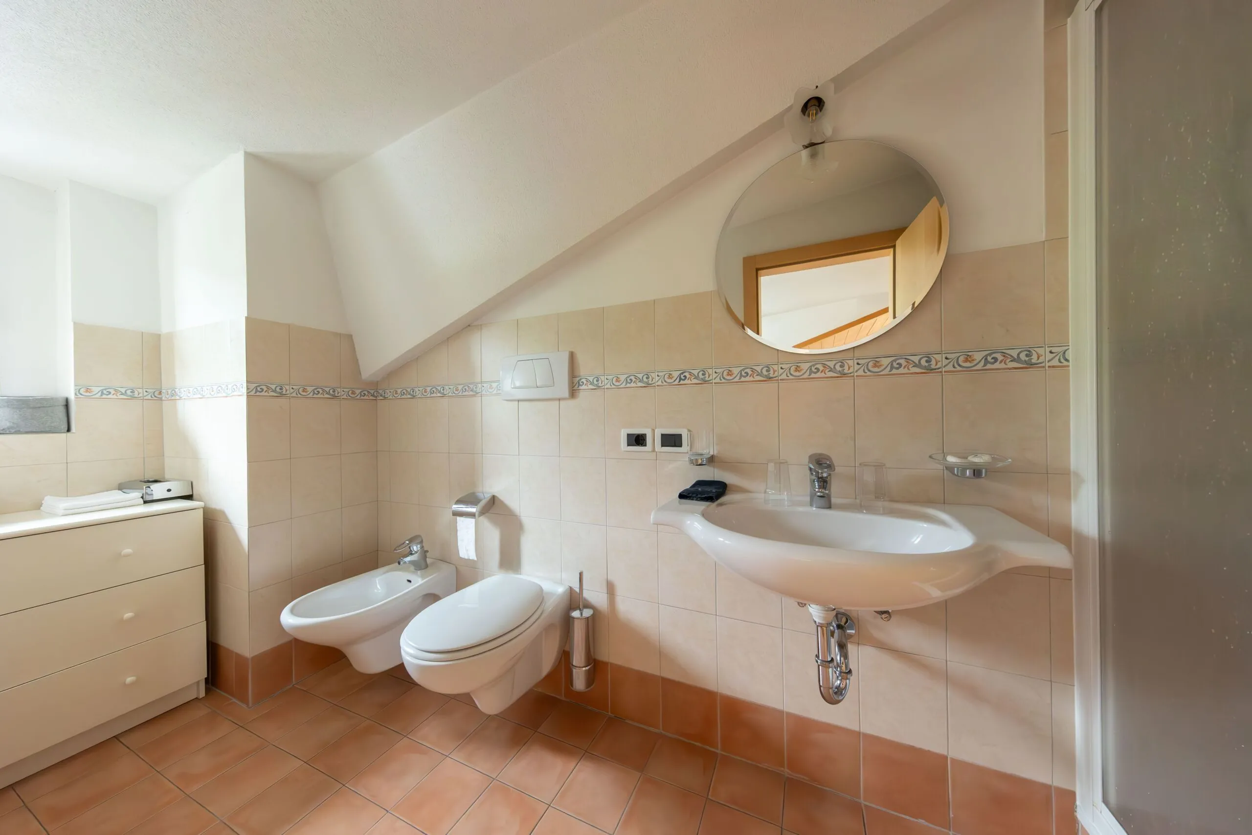 Bright beige-tiled bathroom with wall-mounted sink, bidet, toilet, and round mirror on sloped ceiling wall.