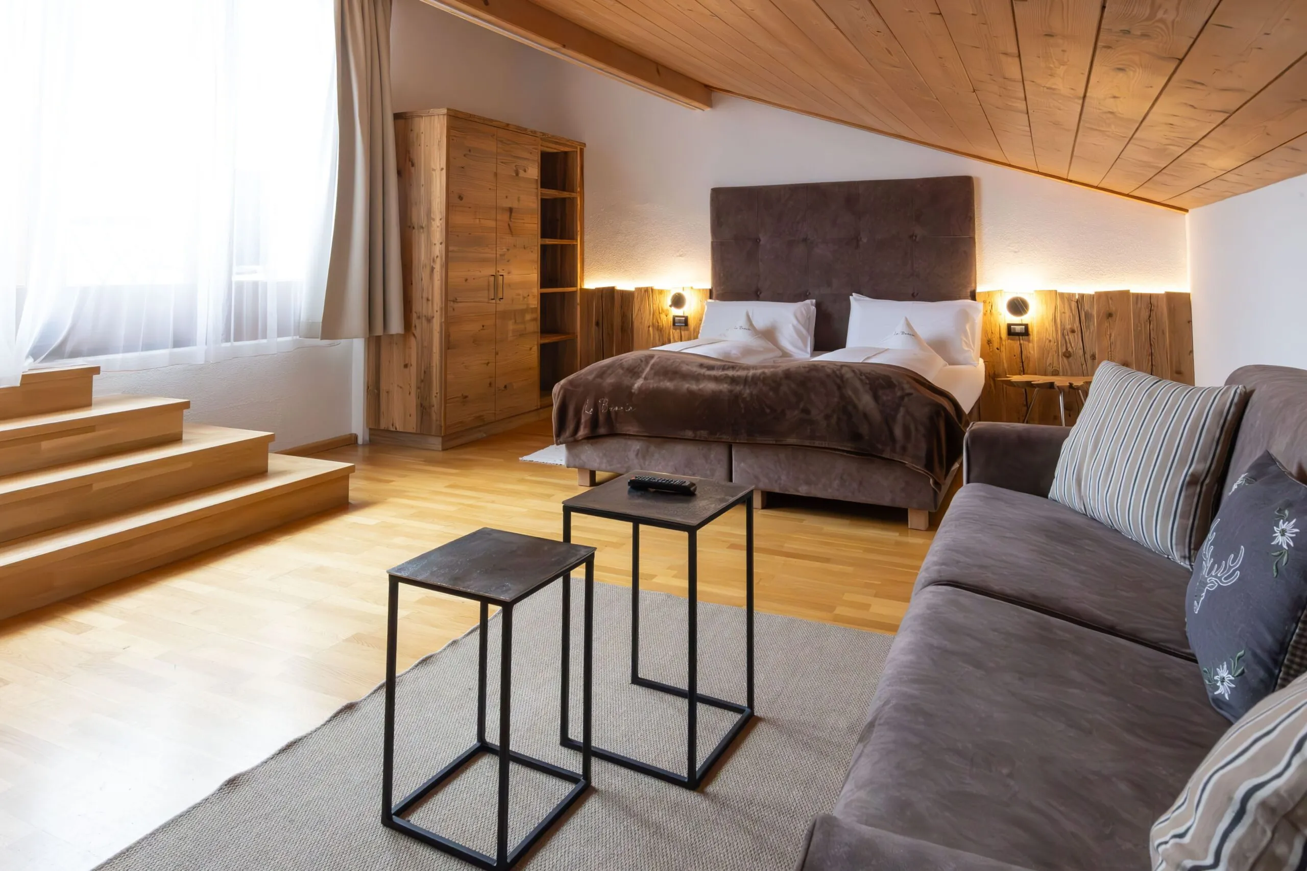Bright hotel suite with king bed, wooden ceiling, warm lighting, and a gray sofa in a cozy Alpine room.