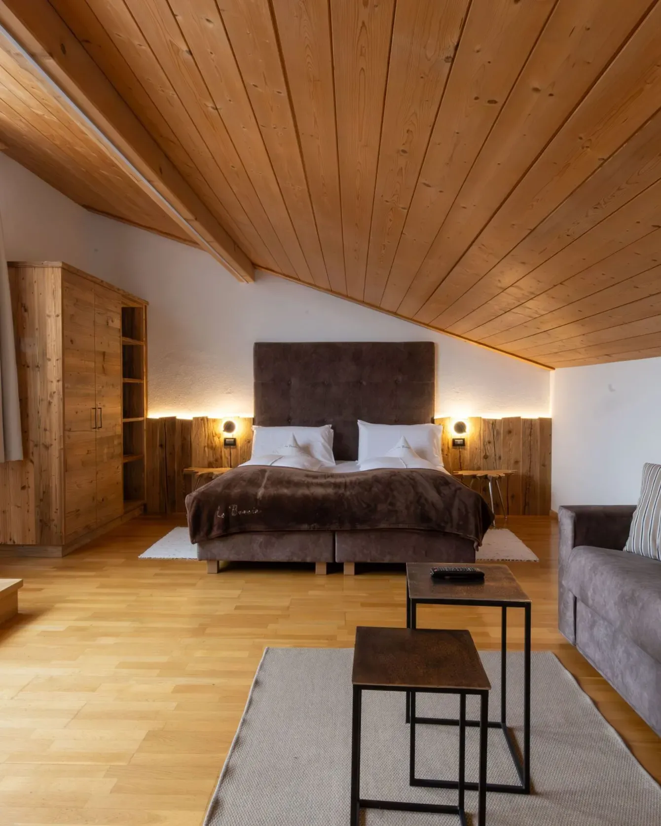 Cozy attic bedroom with a large bed, wooden ceiling, warm lighting, and a gray sofa in front of a small coffee table.