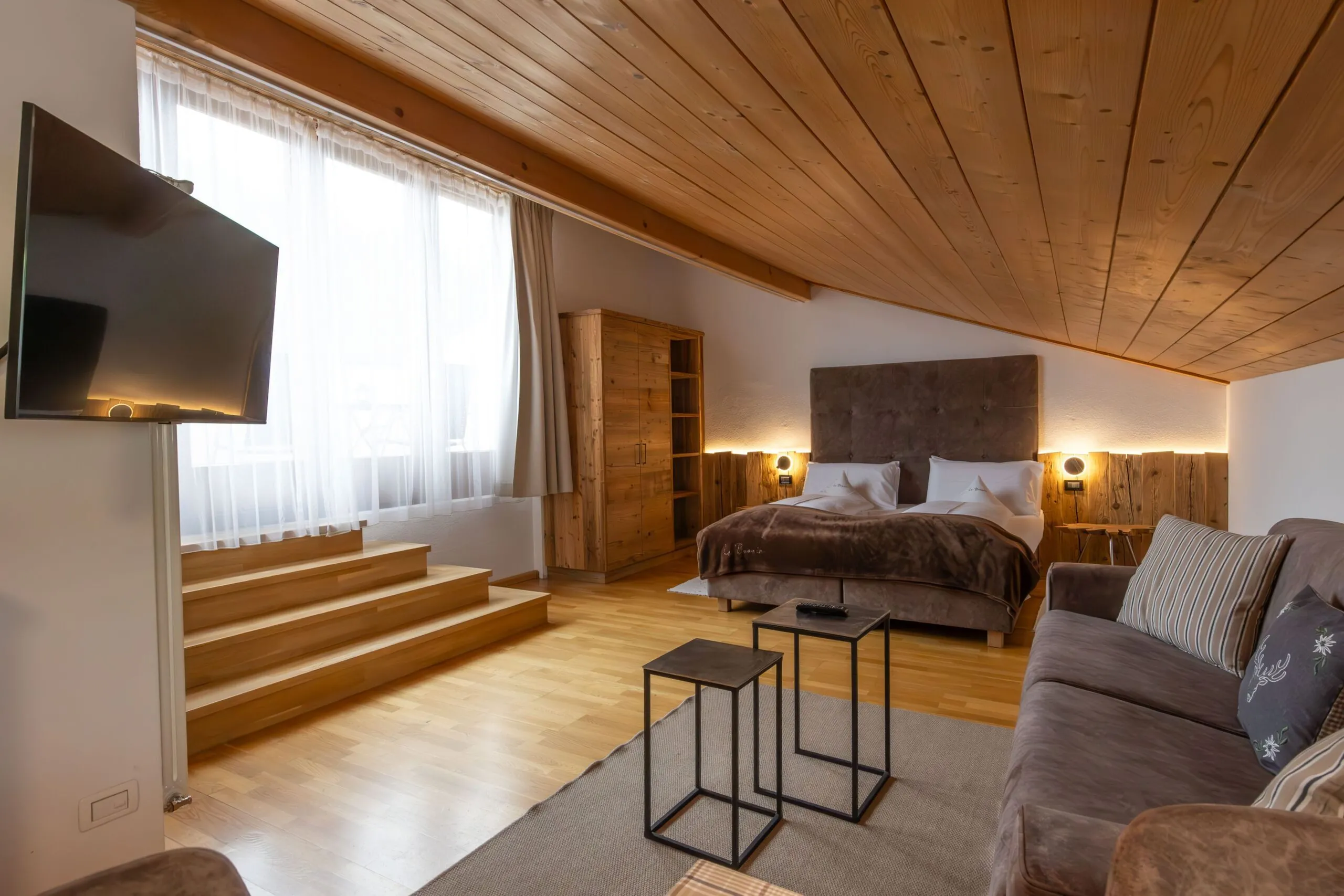 Cozy hotel loft bedroom with wooden ceiling, large bed, TV on wall, and sitting area.