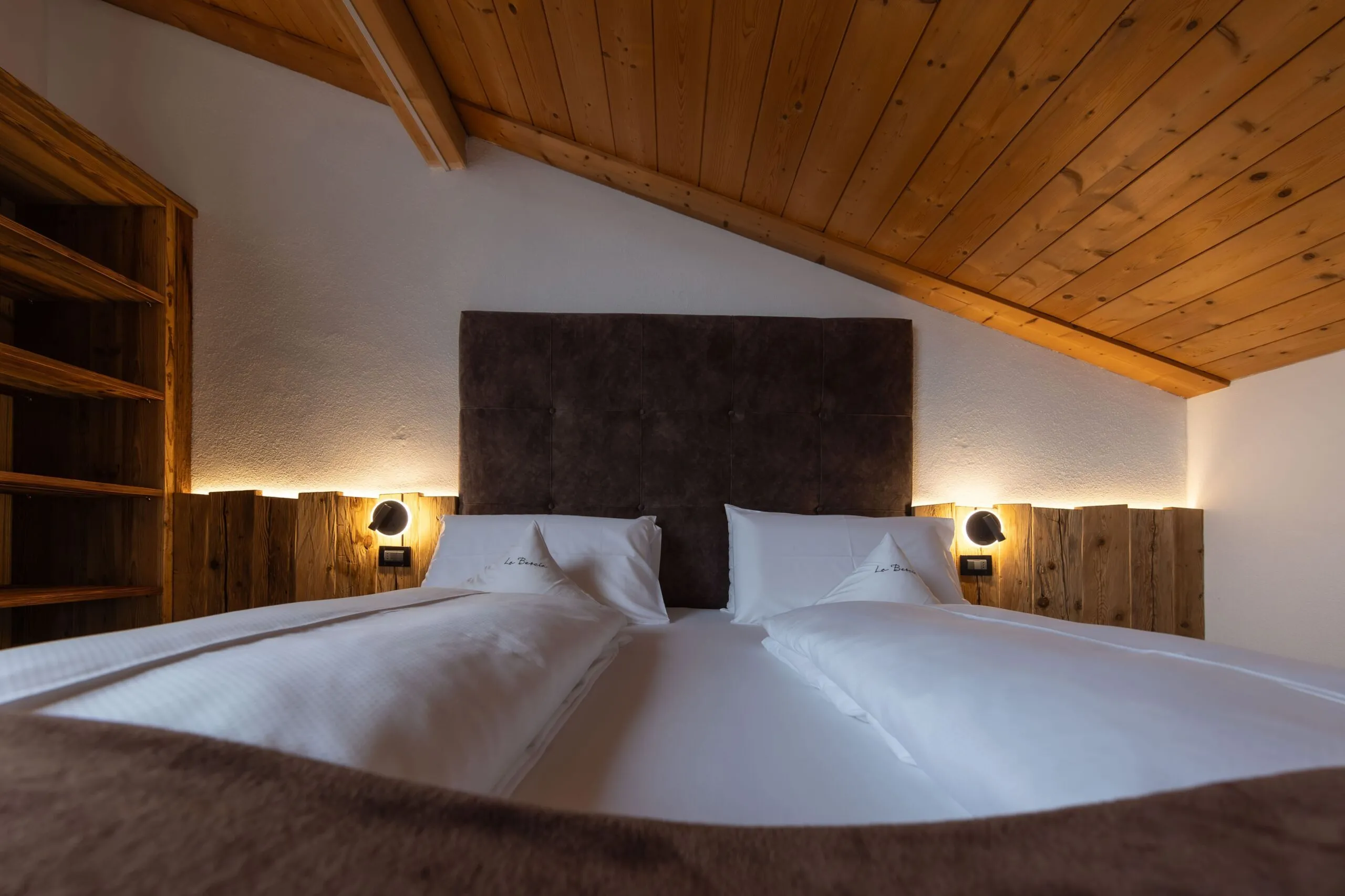 Cozy hotel loft bedroom with a large padded headboard, two white beds, wooden ceiling, and warm wall lights.