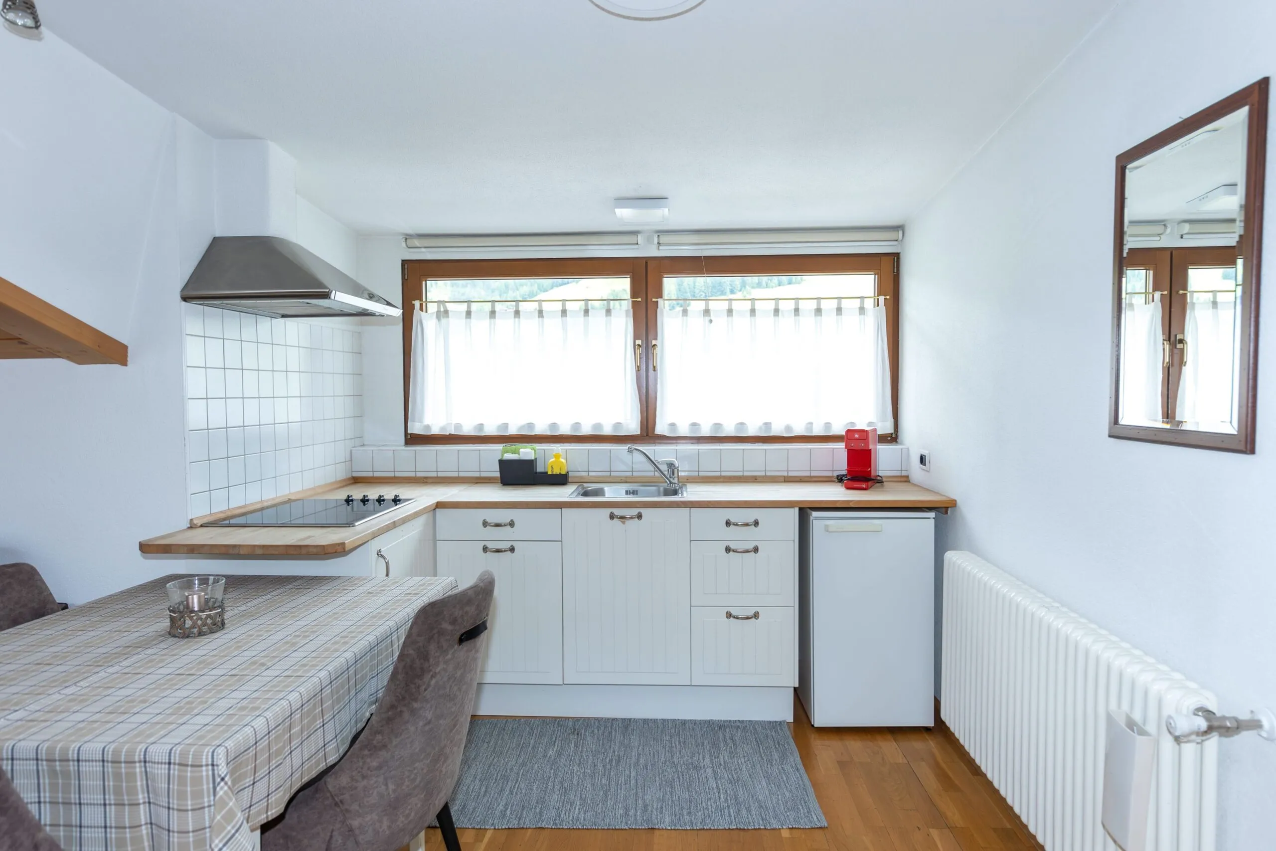 Cozy white kitchen with wooden accents, stove, sink, microwave, red coffee machine, and dining table run along windowed wall.